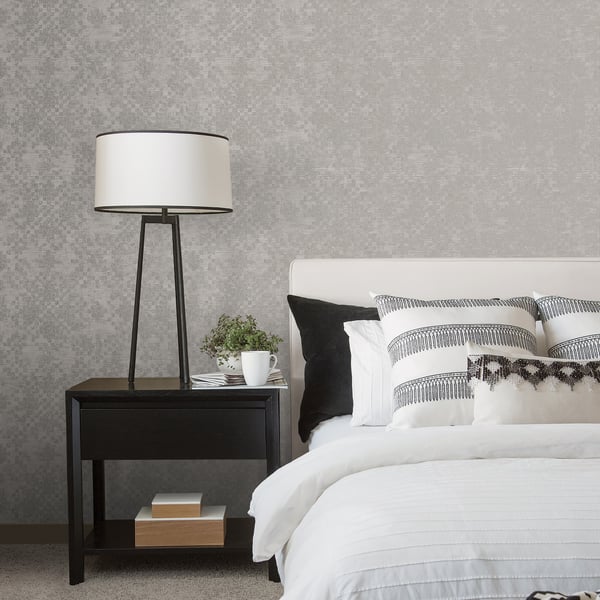 Bedroom wall with silver-grey mosaic tile wallpaper; black tripod bedside lamp, dark nightstand, white bedding, striped cushions.