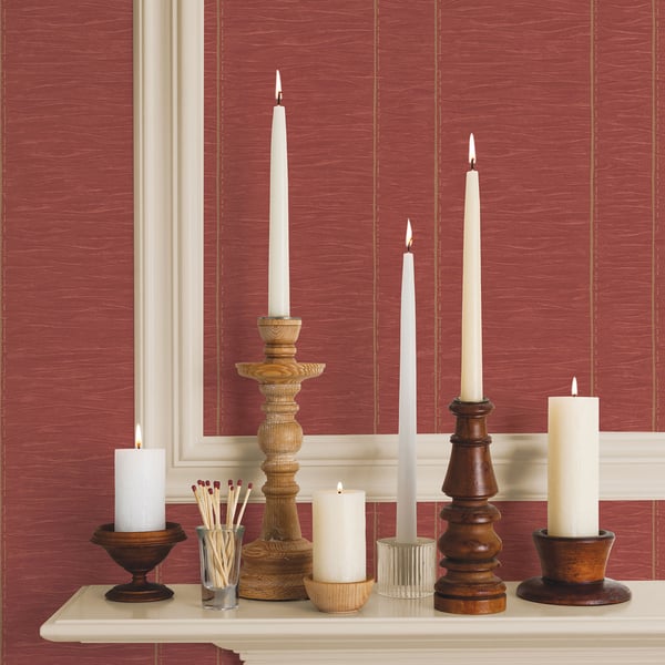 White candles in varying-height wooden and metal candlesticks on a cream mantel with a matches jar, red wallpaper.