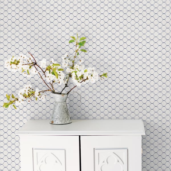 Blue geometric wallpaper backdrop behind a white two-door cabinet topped by a metal pitcher vase with white blossoms and branches.