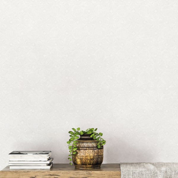 White wallpaper backdrop with a gold-toned ceramic vase featuring geometric patterns, a trailing green plant, and a small stack of books on a wooden shelf.