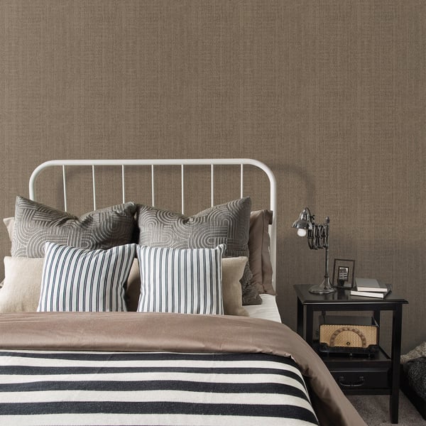 White metal bed frame with striped blue pillows and beige bedding against brown textured wallpaper, black nightstand.