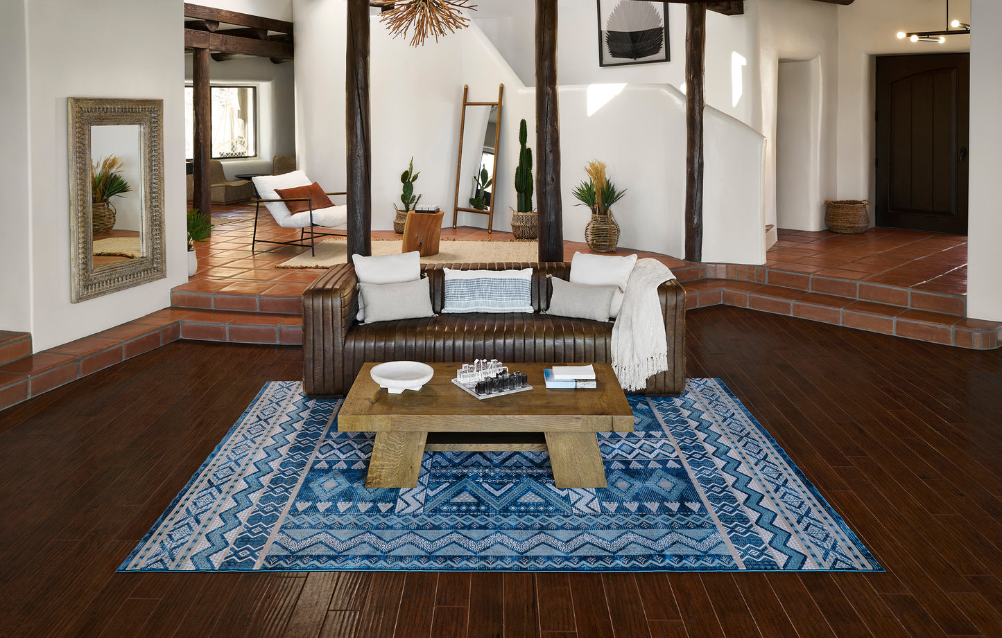 Blue geometric rug in a bright living room, with a brown leather sofa, white and blue cushions, and a rustic wooden coffee table.