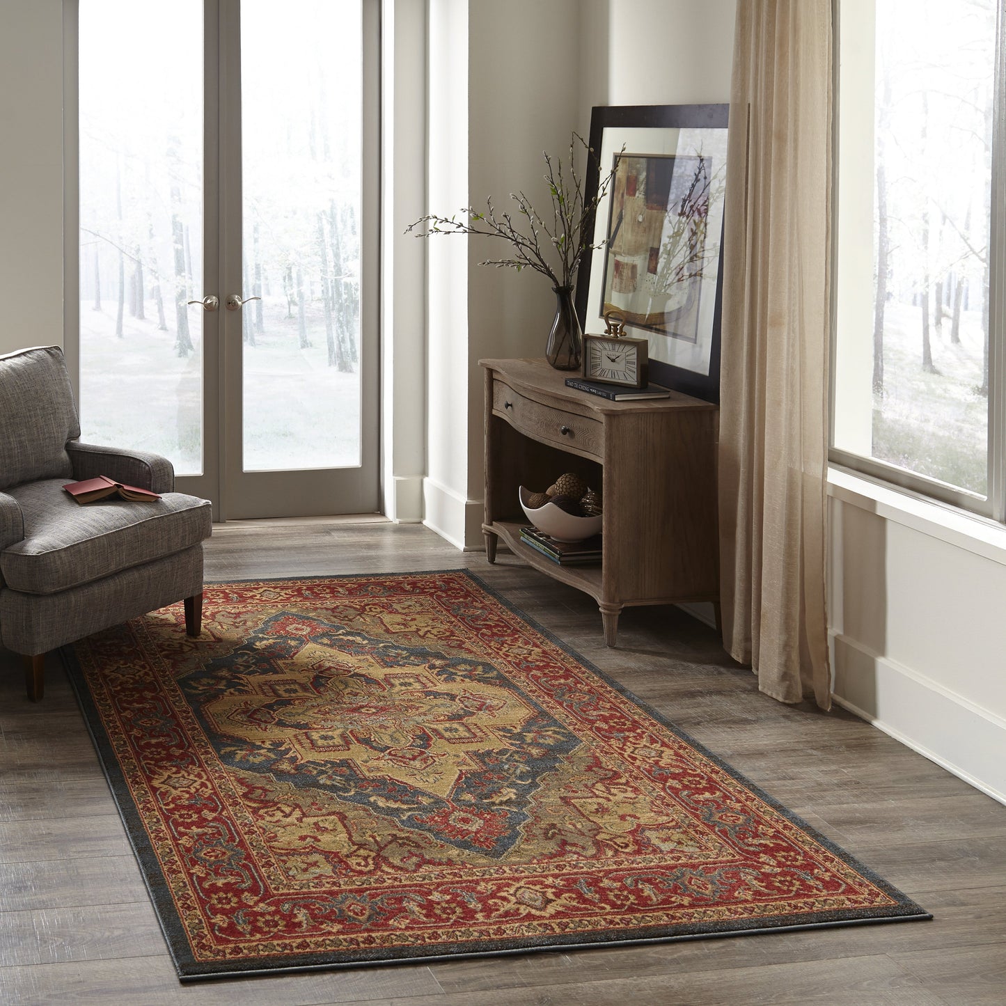 Oriental area rug in red, blue and gold with a dark border, on wood flooring beside a grey armchair and wooden console table.