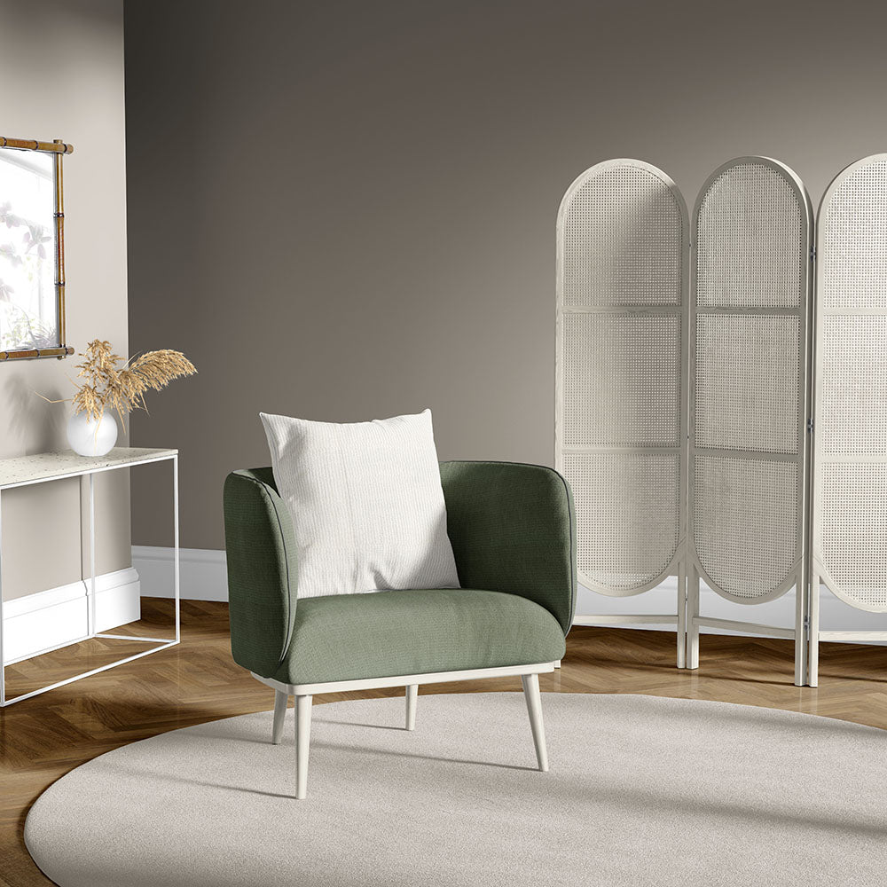 Olive green fabric armchair with light wood legs and white pillow, in a neutral living room with a woven room divider.
