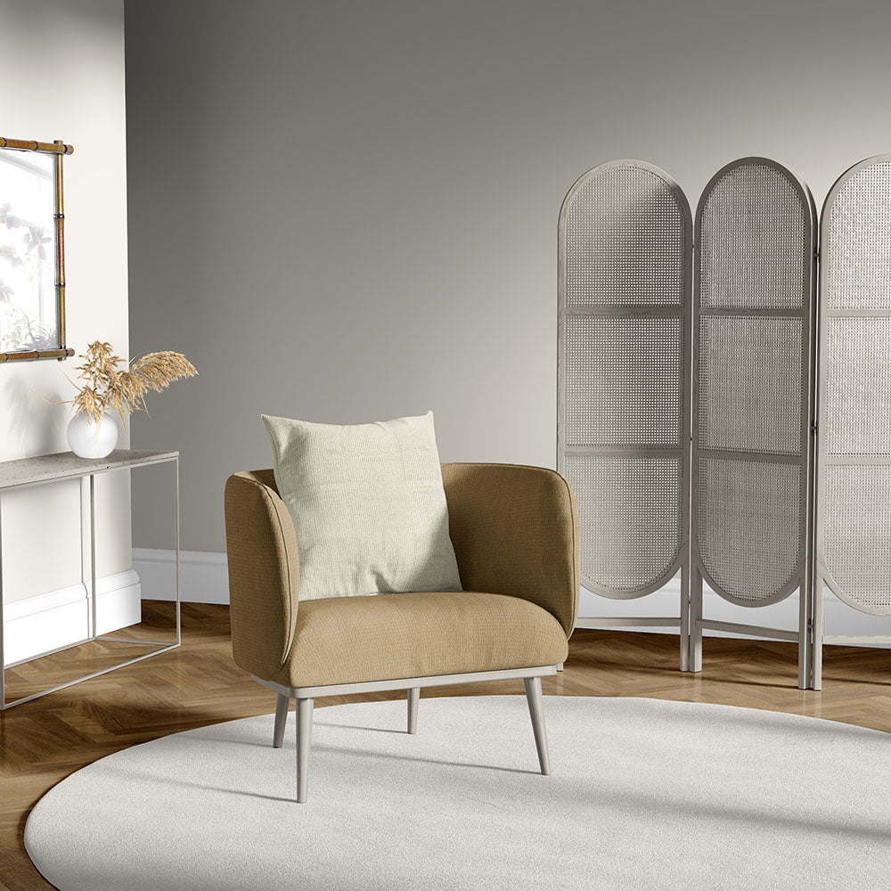 Beige upholstered armchair with pale cushion, light round rug, woven rattan room divider, and slim console table with a vase of dried stems.