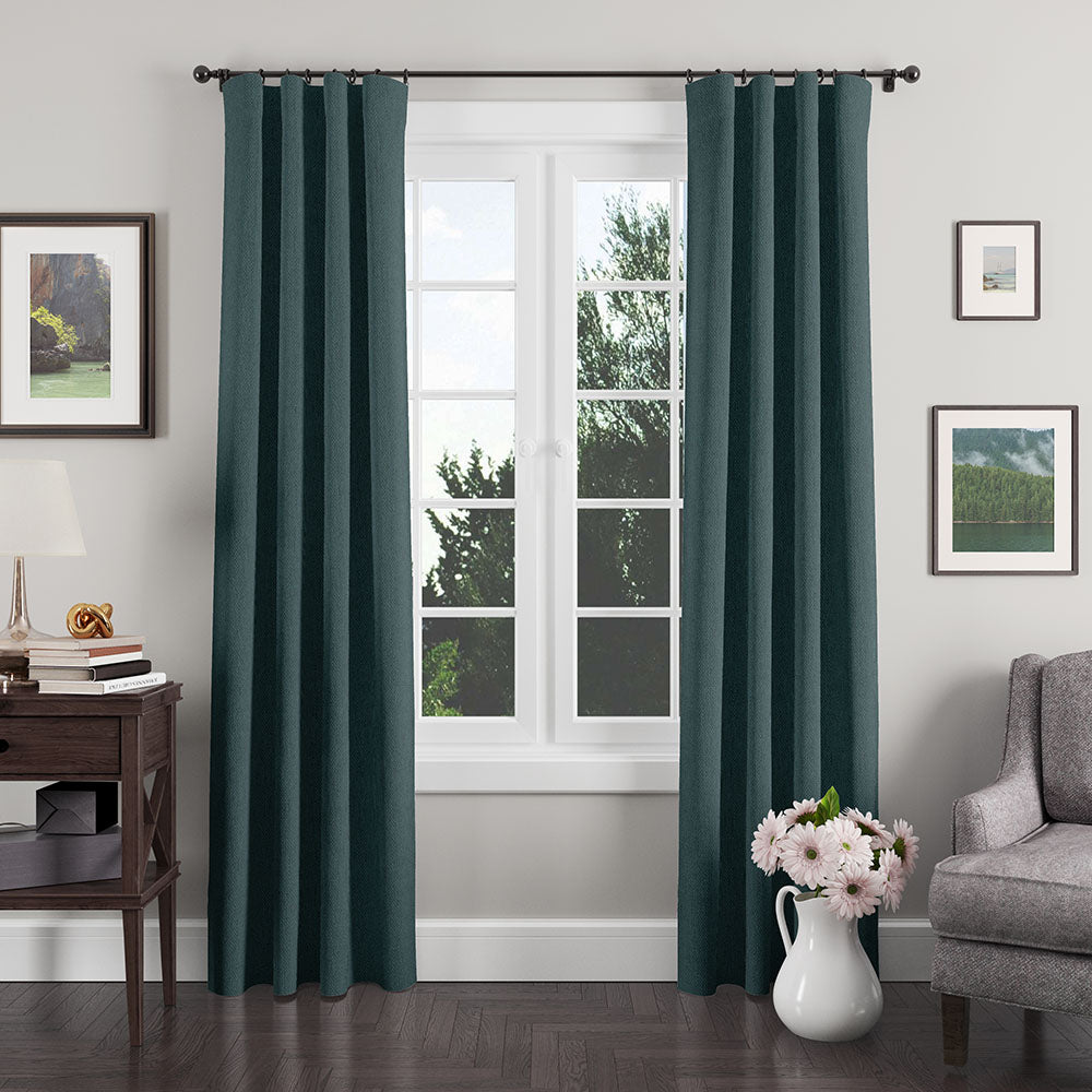 Teal curtains frame a white window in a cozy living room; gray armchair, dark wood side table, lamp, & vase of pink flowers.