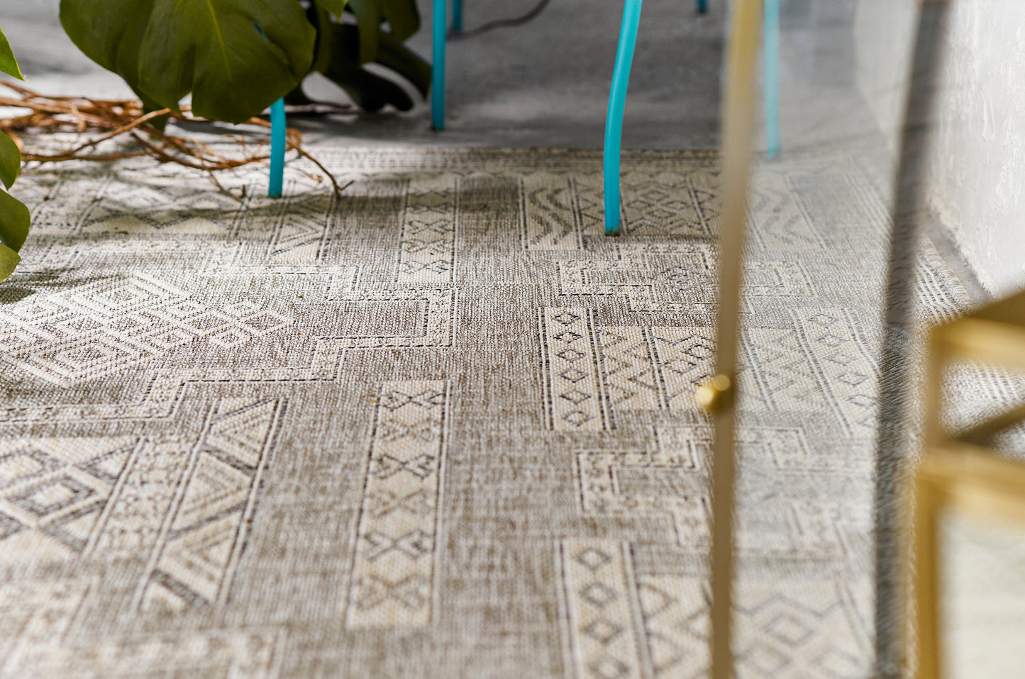 Beige-gray woven geometric rug with tribal motifs, turquoise chair legs, and a gold furniture frame.