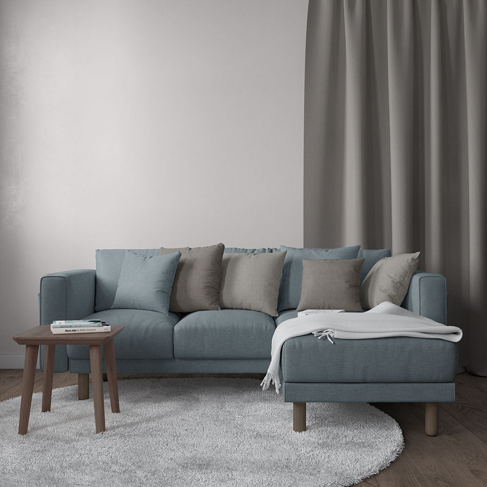 Blue-gray fabric sectional sofa with chaise, blue and taupe cushions, throw blanket, wooden legs; side table, gray rug, gray curtains.