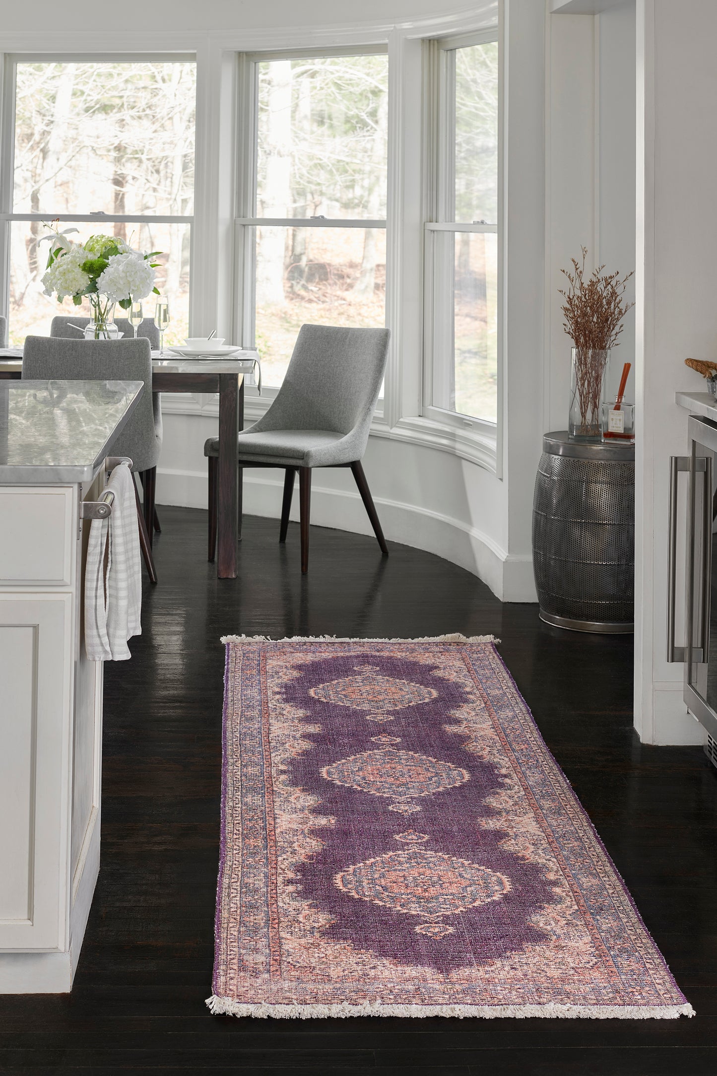 Purple Persian-inspired runner rug with ivory fringe on dark wood floor beside white kitchen and grey dining chairs.