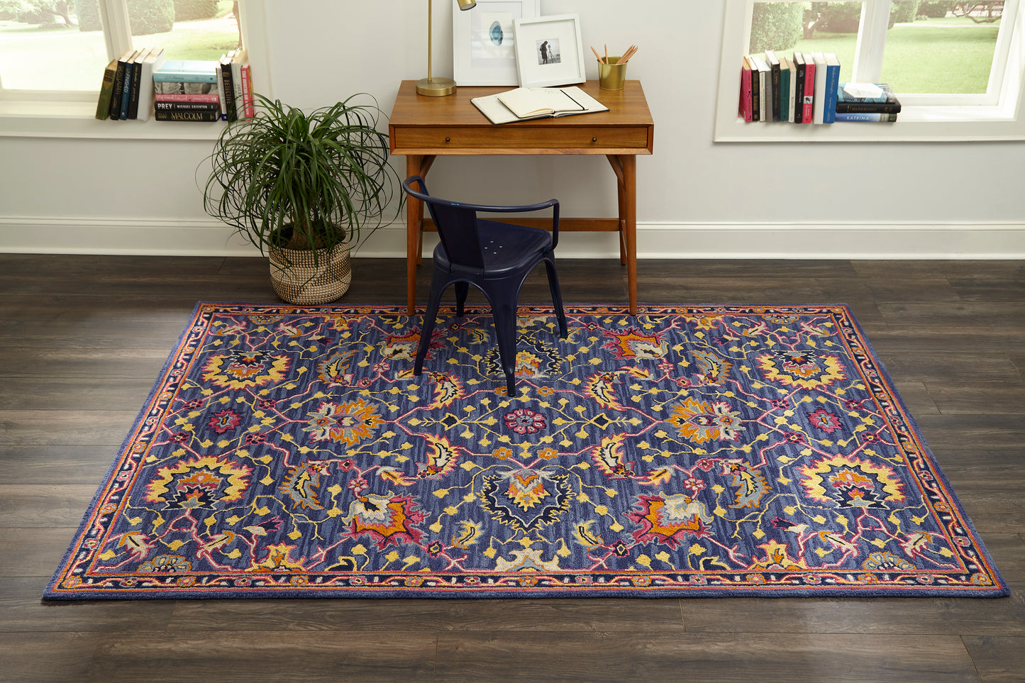 Blue oriental rug with multicolor floral motifs and orange border in a home office with a wooden desk and navy chair.