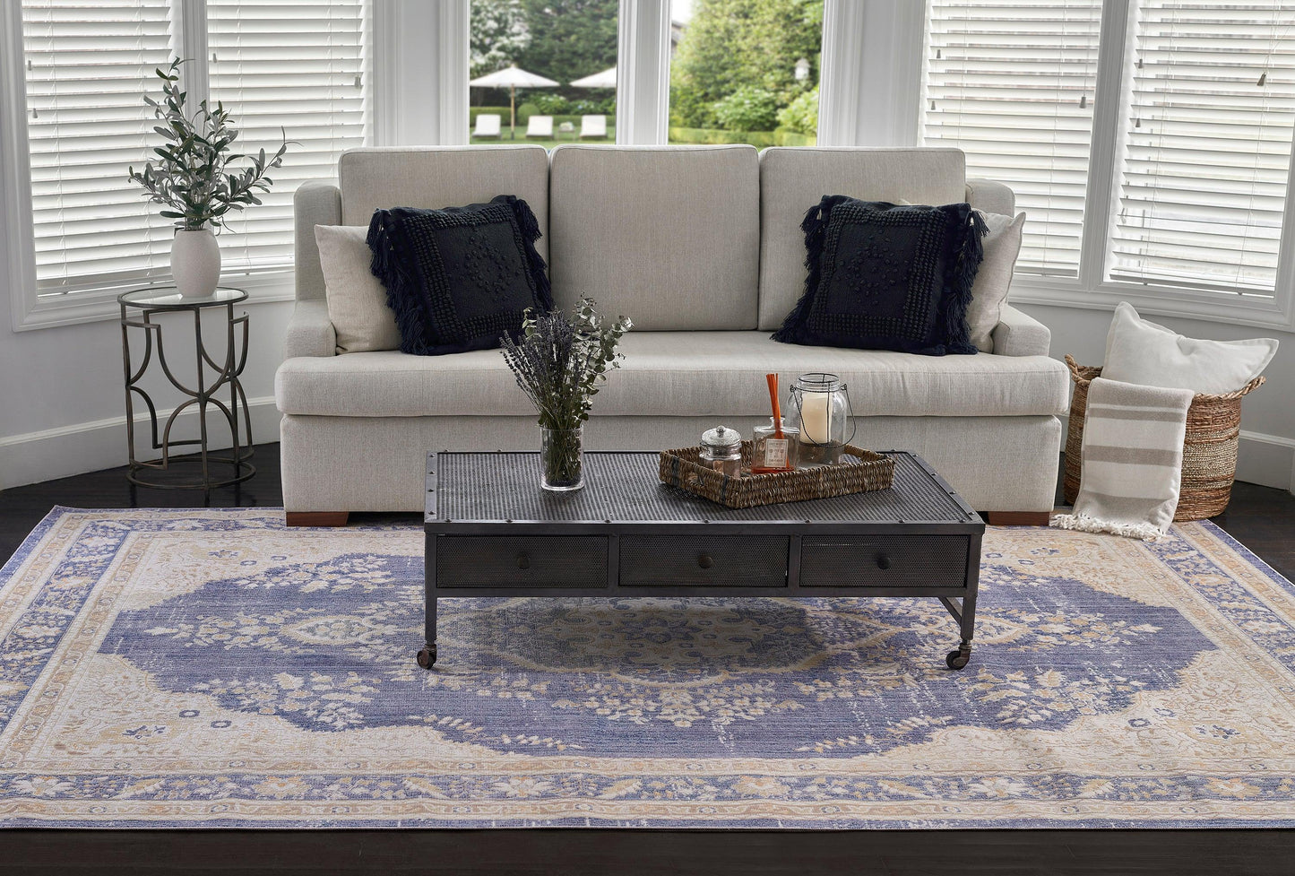 Cream sofa with black cushions, dark metal coffee table with drawers, blue and ivory ornate rug, wicker basket and greenery.