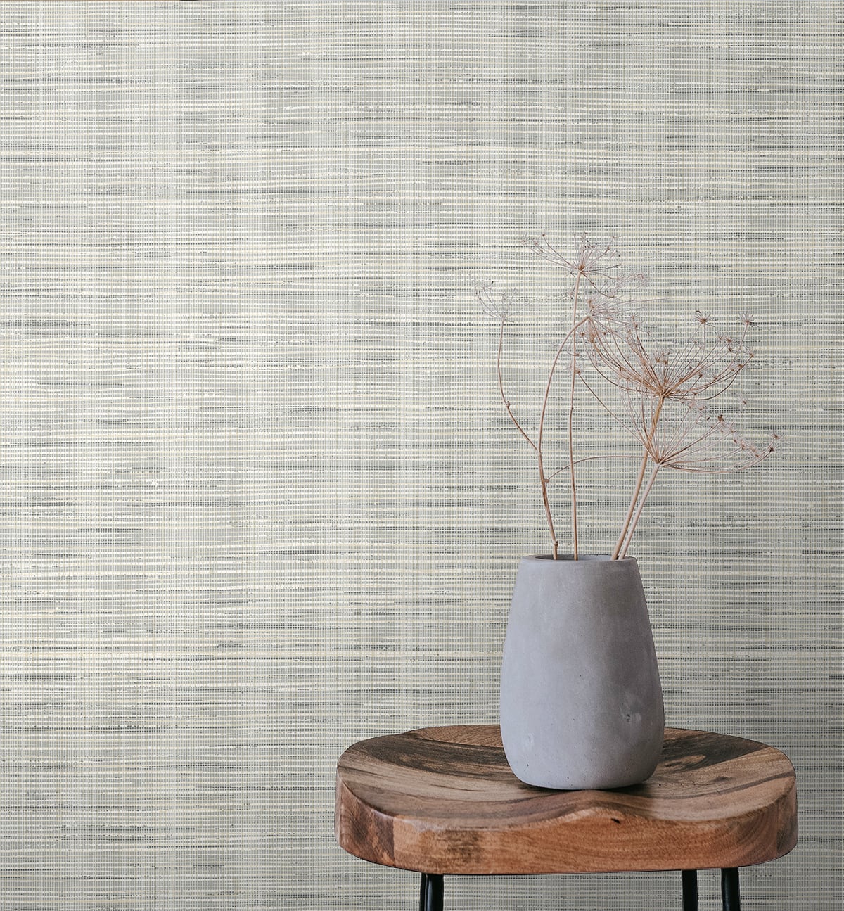 Gray ceramic vase with dried delicate stems on a round wooden slab against a textured beige wall.