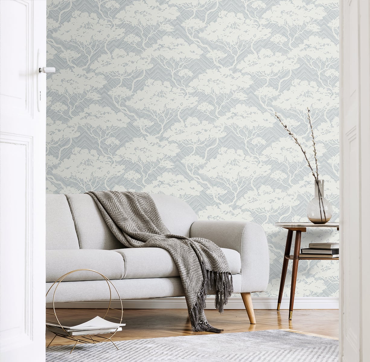 Light gray sofa with a knit throw in a blue-gray botanical wallpaper living room; wooden side table with a vase.