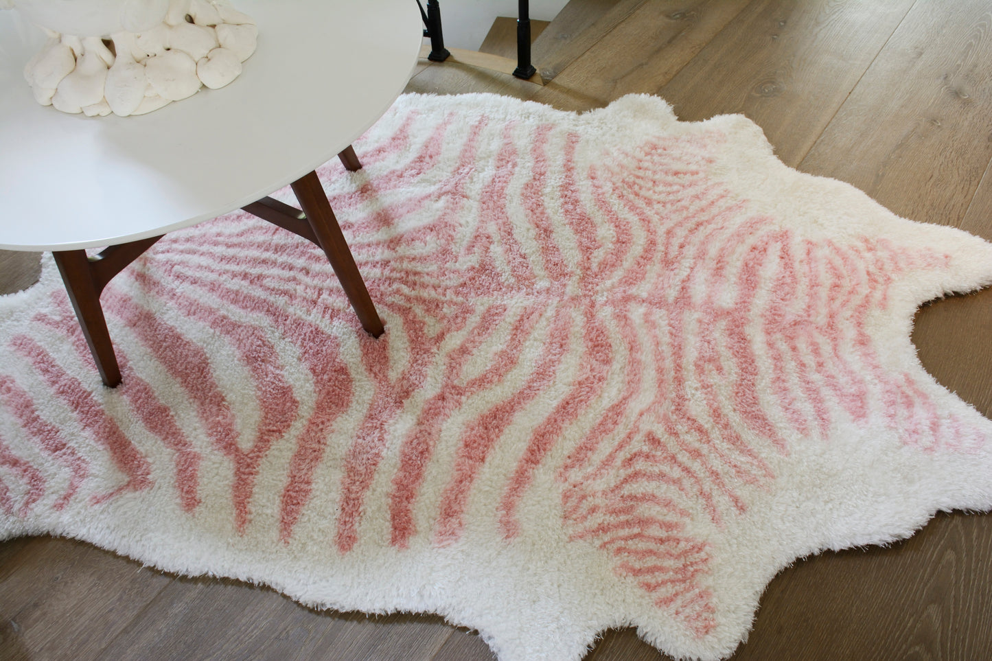 Fluffy pink zebra-print shag rug with white edges, irregular shape on wood floor under a white round table.