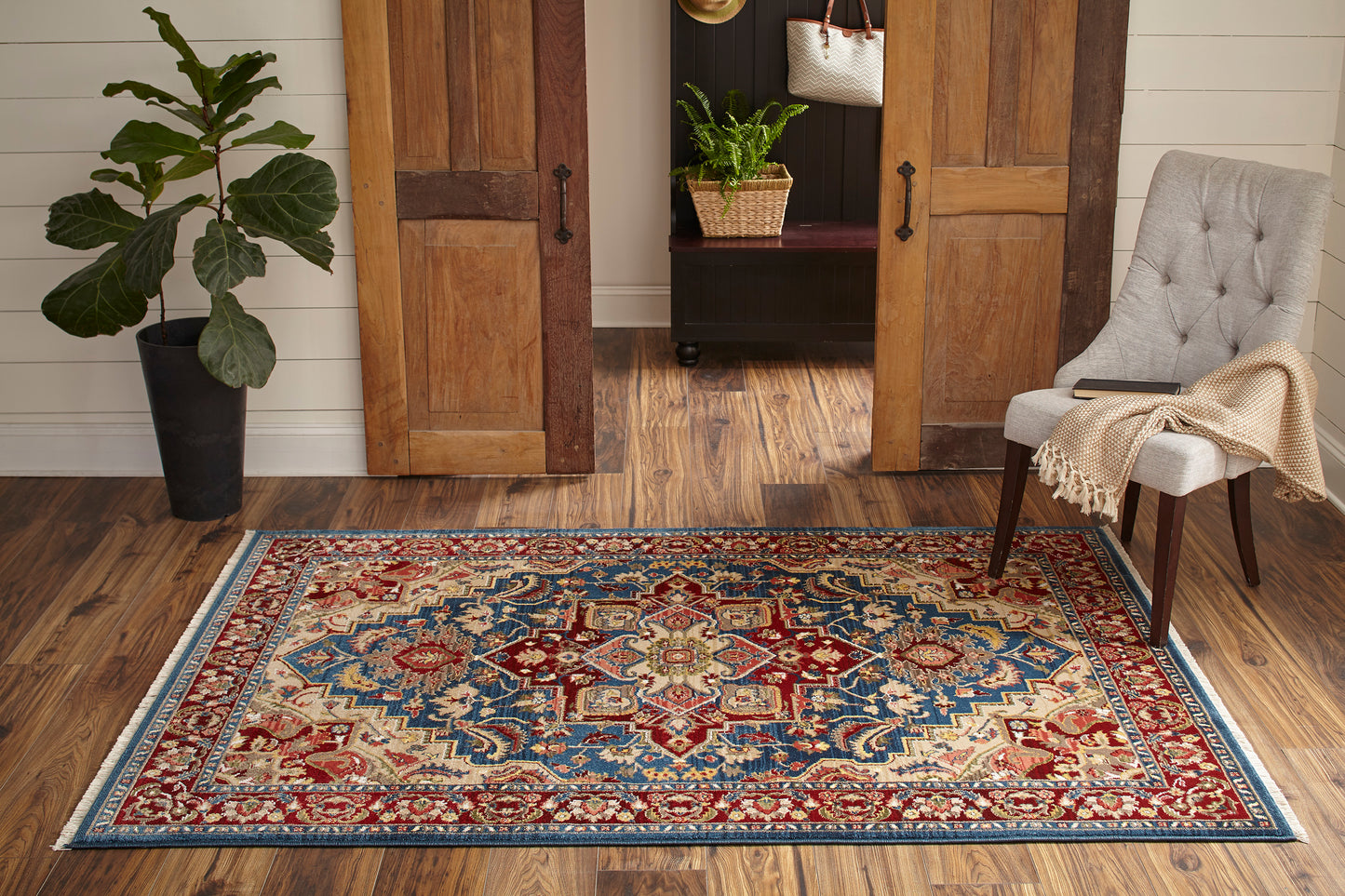 Blue oriental area rug with red and ivory motifs on a wood floor, flanked by a plant and a tufted chair.