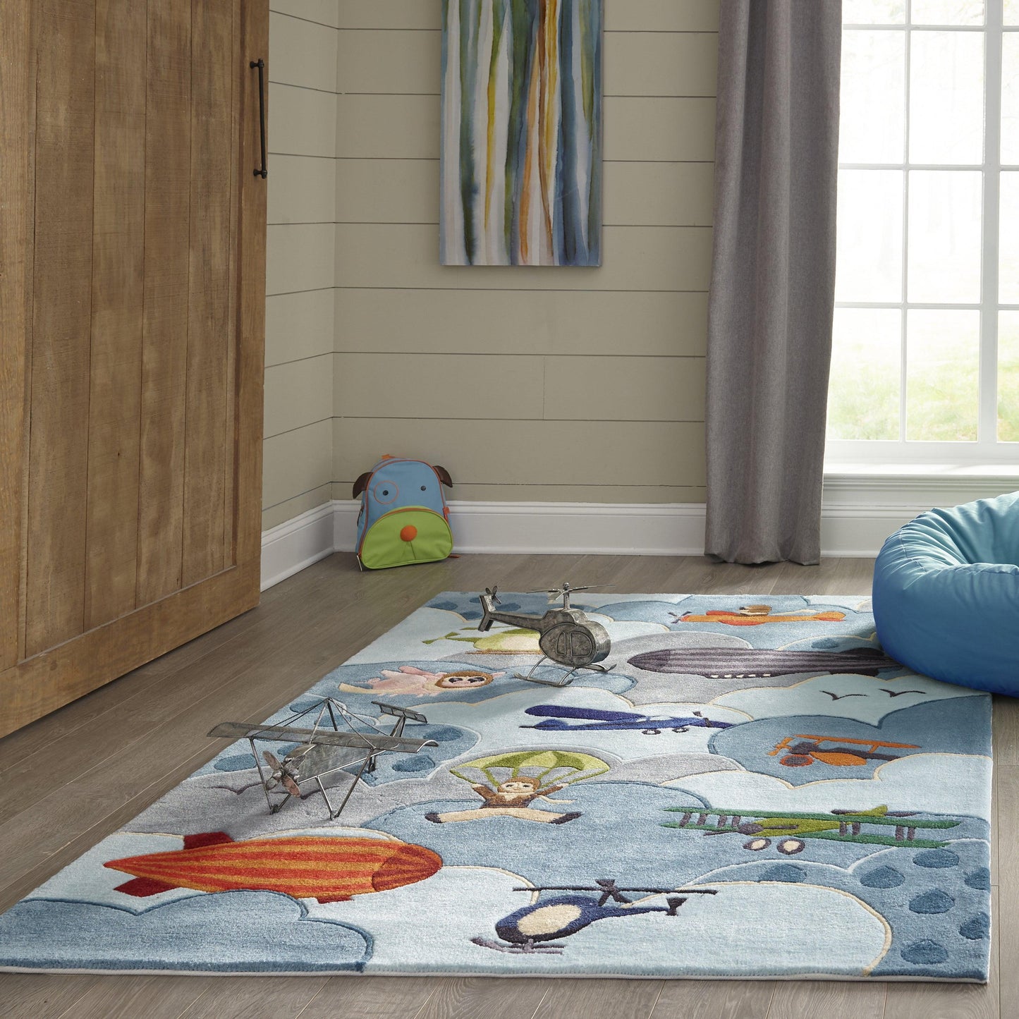 Rectangular kids' rug in sky blue with airplanes and clouds; cozy playroom scene with bean bag and wooden cabinet.