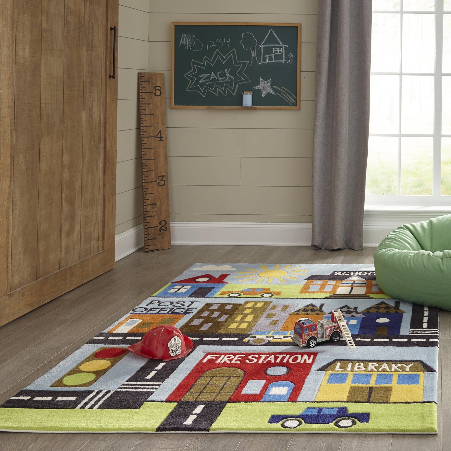 Colorful kids' city rug (5' round) with roads, buildings, a fire station and library; ruler and a red cap in a cozy playroom.