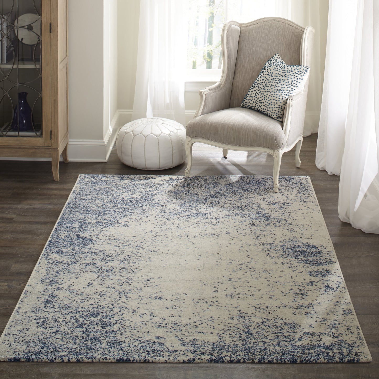 Blue and ivory distressed-pattern rectangular rug, low-pile, in a light-filled living room with a beige wingback chair and white pouf.