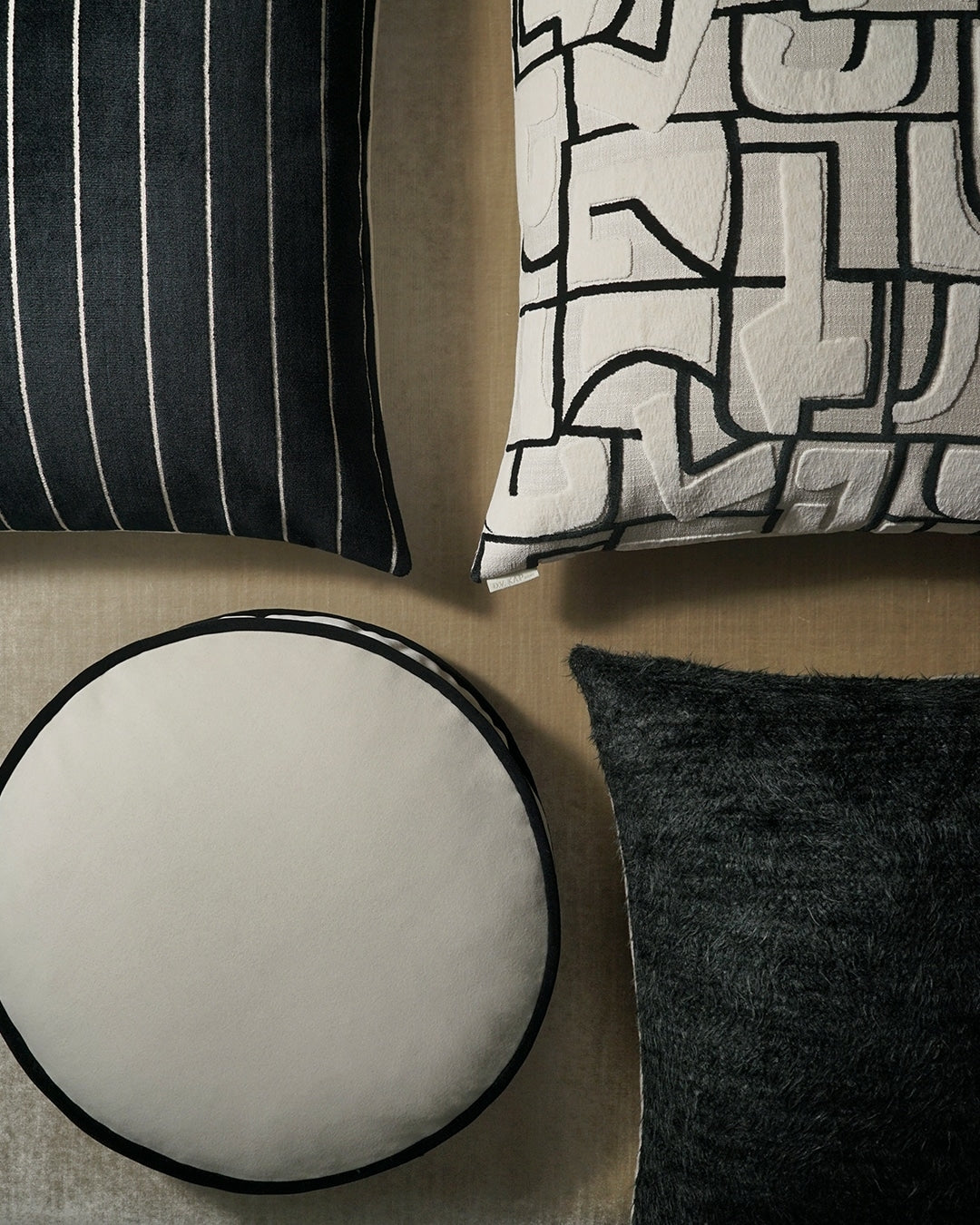 Decorative cushions: black striped, ivory geometric, and dark charcoal textures; round ivory pouf on beige surface.