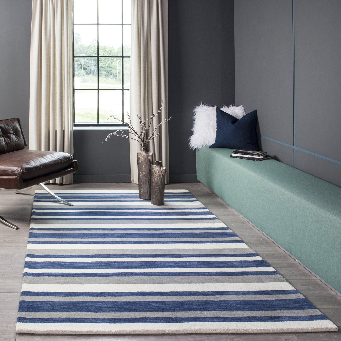 Modern living room with a blue and white striped rug, teal bench, navy cushion, fluffy white pillow, brown leather chair.