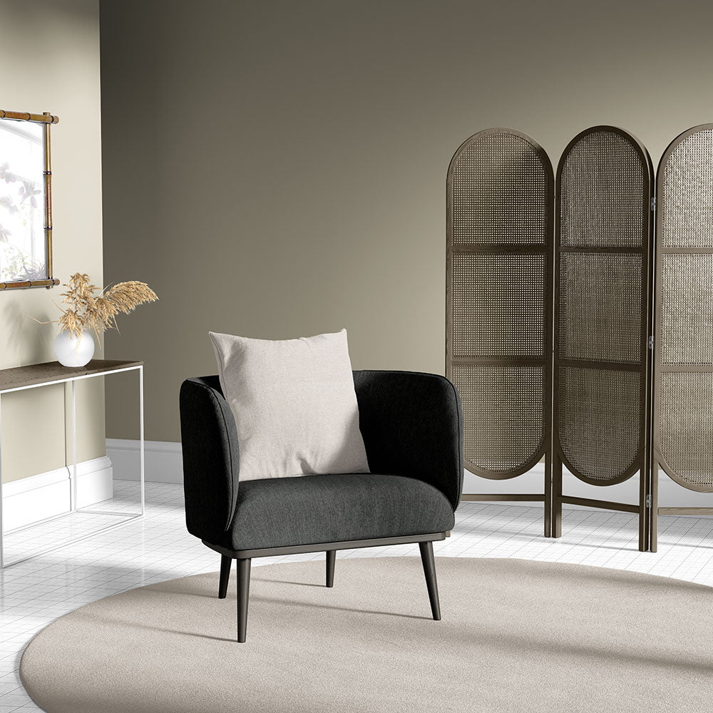Charcoal gray mid-century armchair with a light beige cushion on a round beige rug beside a wicker folding screen.