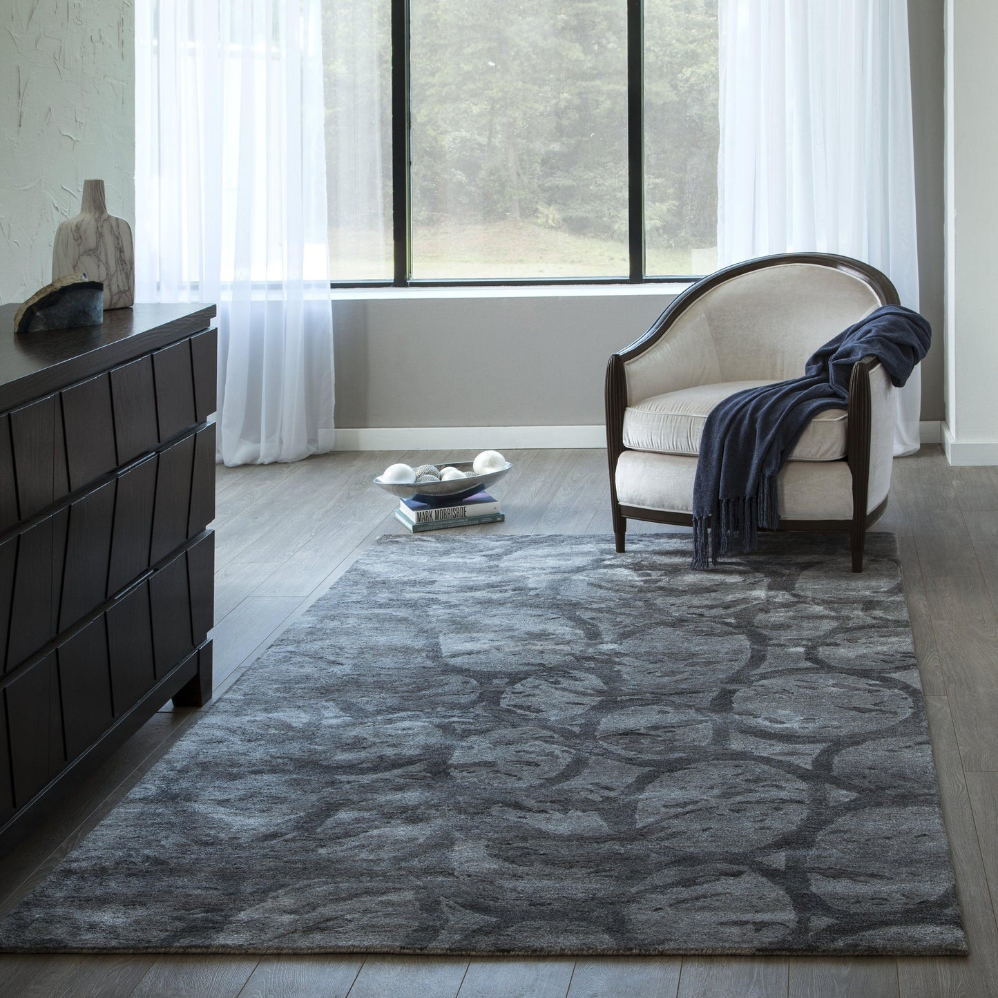 Grey patterned area rug in modern living room with dark wood dresser, beige upholstered armchair, navy throw, large window.