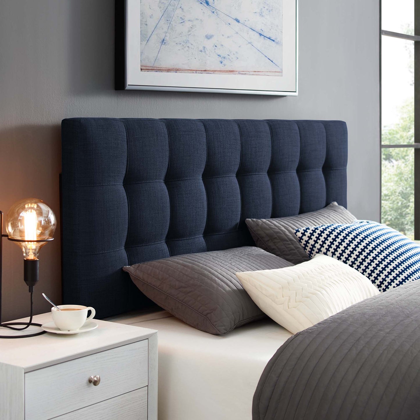 Navy tufted headboard with gray quilted bedding, white patterned cushions, white nightstand, Edison lamp.