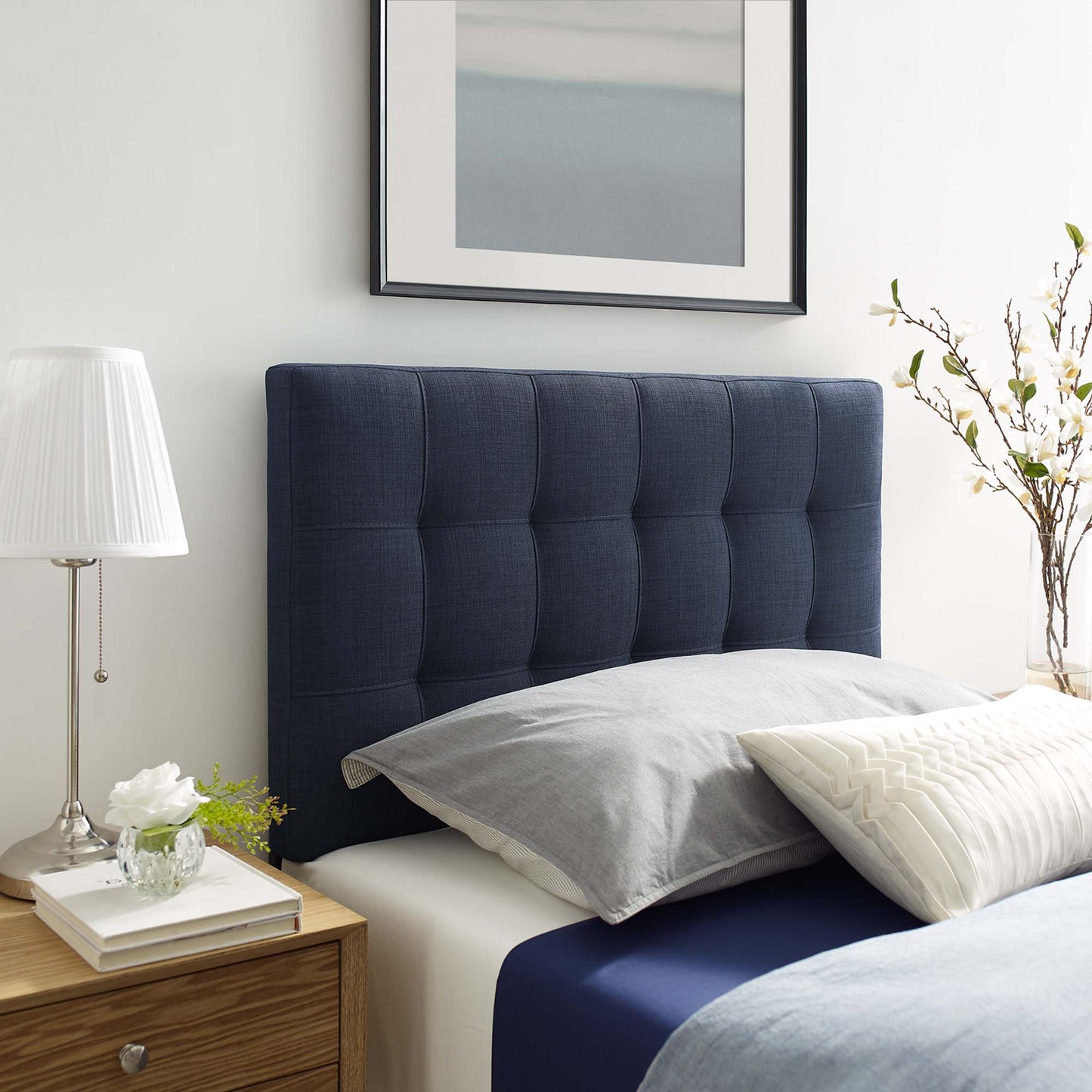 Navy tufted upholstered twin headboard with gray pillows and navy sheets; wooden nightstand, lamp & framed art.