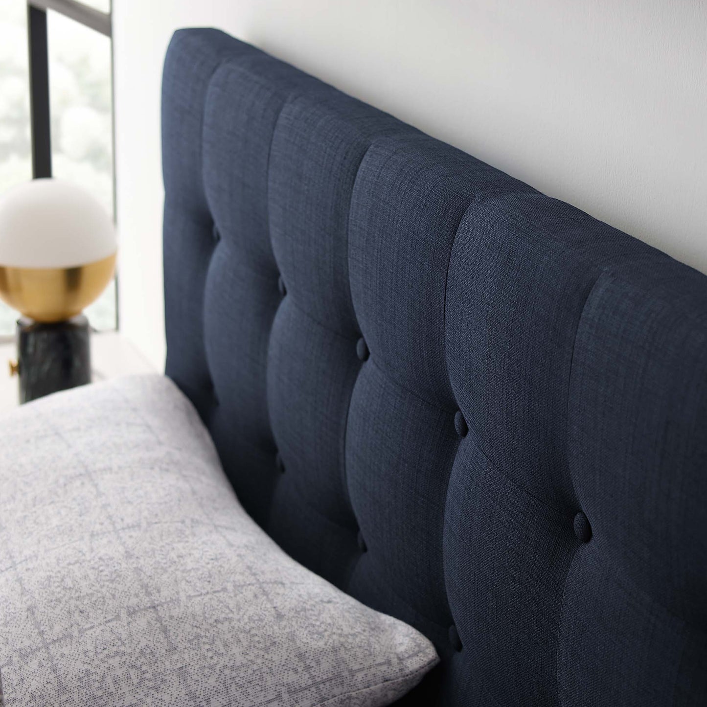 Navy blue tufted fabric headboard with button details beside a light gray quilt and gold bedside lamp.