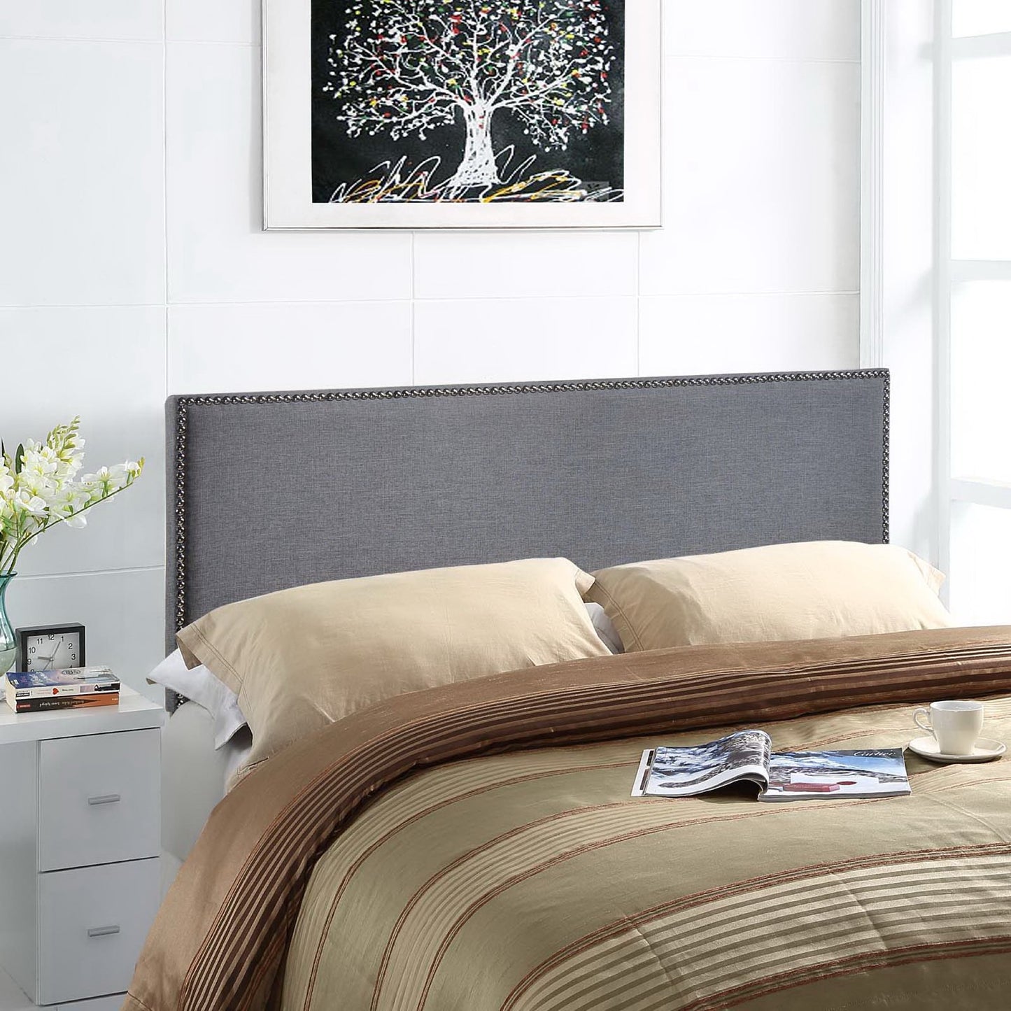 Grey upholstered bed headboard with nailhead trim, beige sheets, brown striped comforter; nightstand, vase of flowers, & tree artwork above.