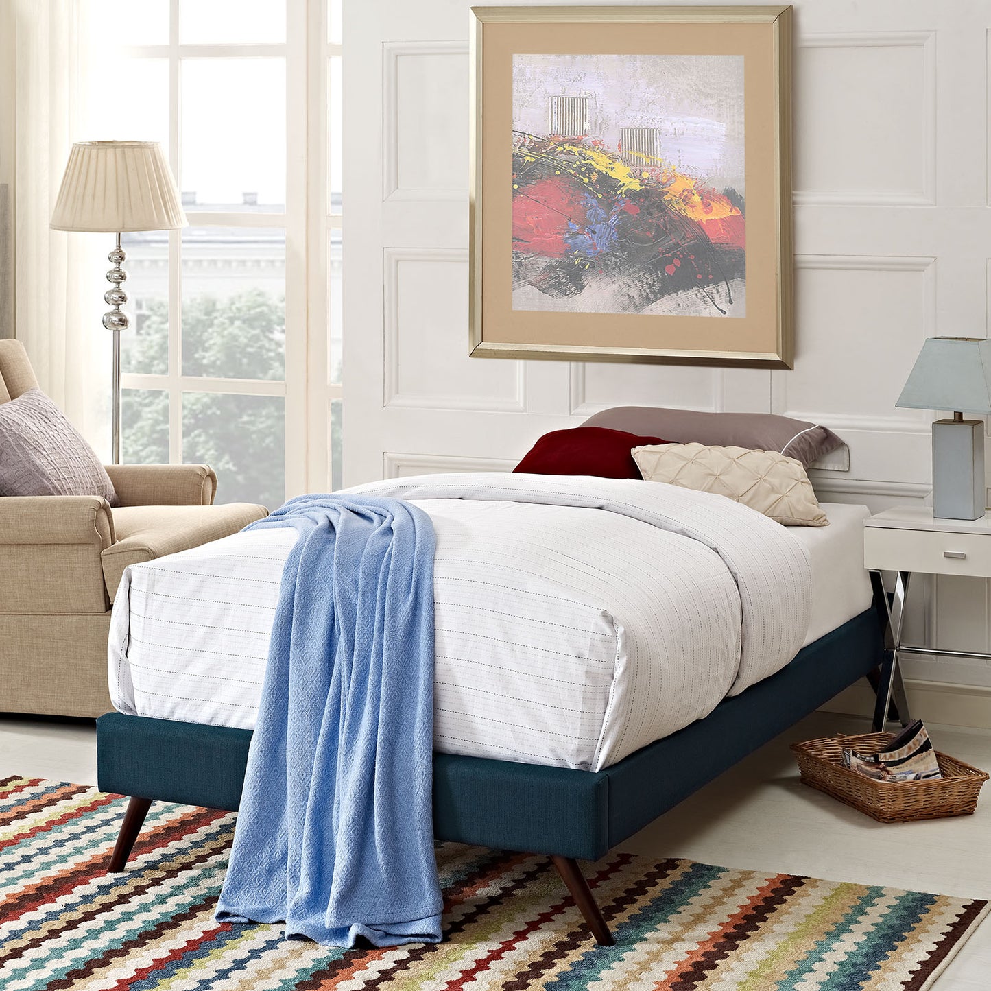 Teal upholstered twin bed frame with white linens and a light blue throw, round splayed legs, beside a multicolor striped rug and a beige chair.