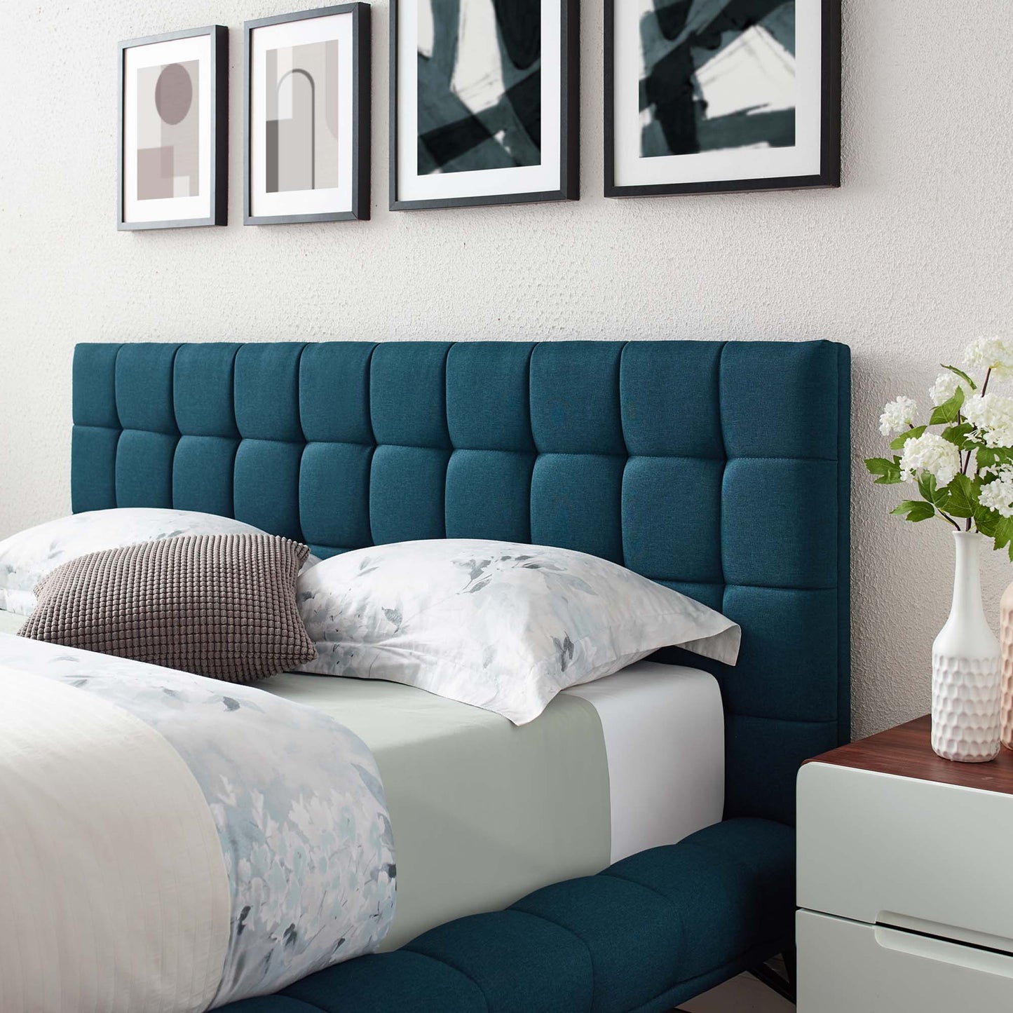 Blue tufted upholstered queen platform bed with square-panel headboard and footboard; white bedding, nightstand, vase of flowers, wall art