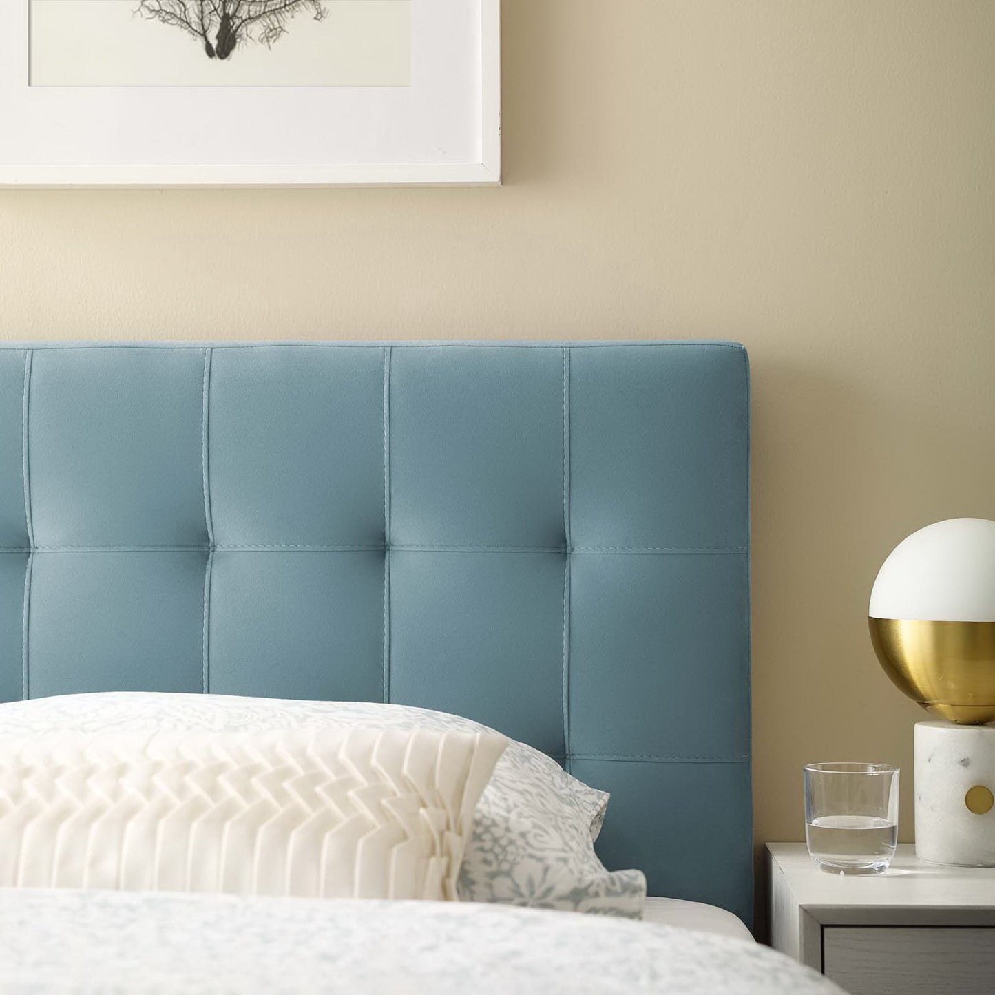 Blue tufted headboard on a bed with cream pillows, beige wall, framed art, and white nightstand with a gold lamp.