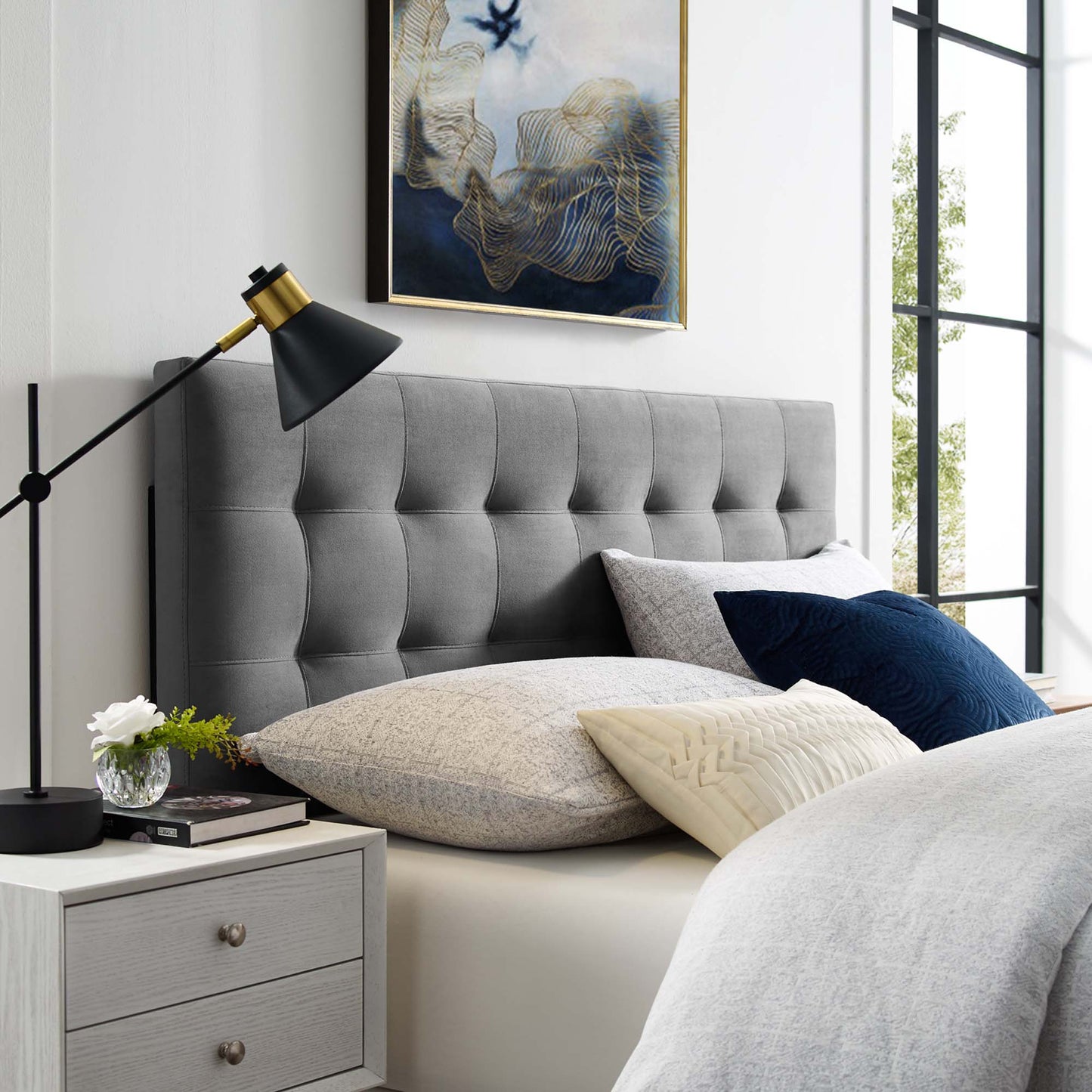 Grey tufted headboard, matching nightstand, black-gold lamp, beige bedding, navy pillow, framed abstract art, large window.