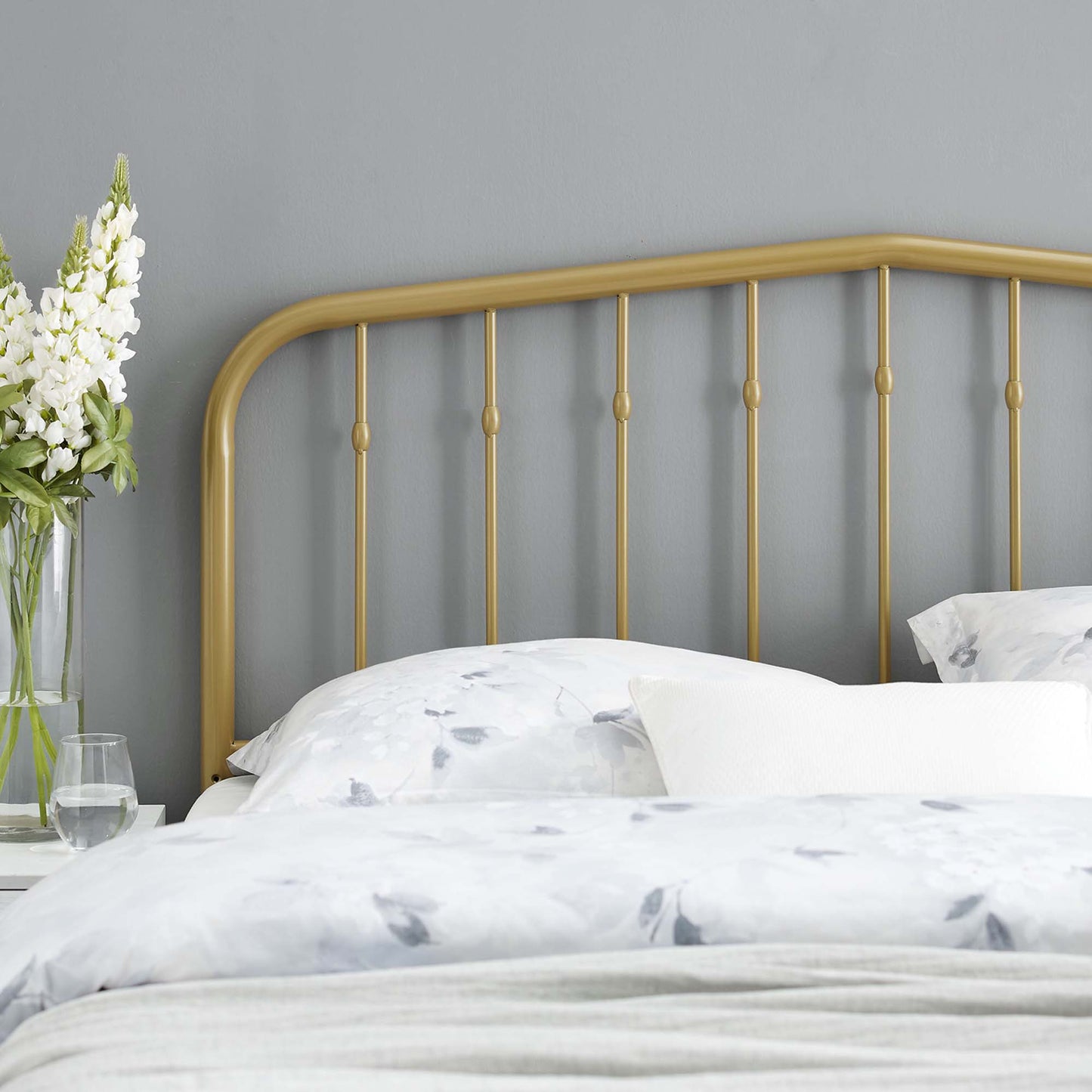 Yellow metal headboard with a curved top rail and vertical posts, white floral bedding, gray wall, and a vase of white flowers on the nightstand.