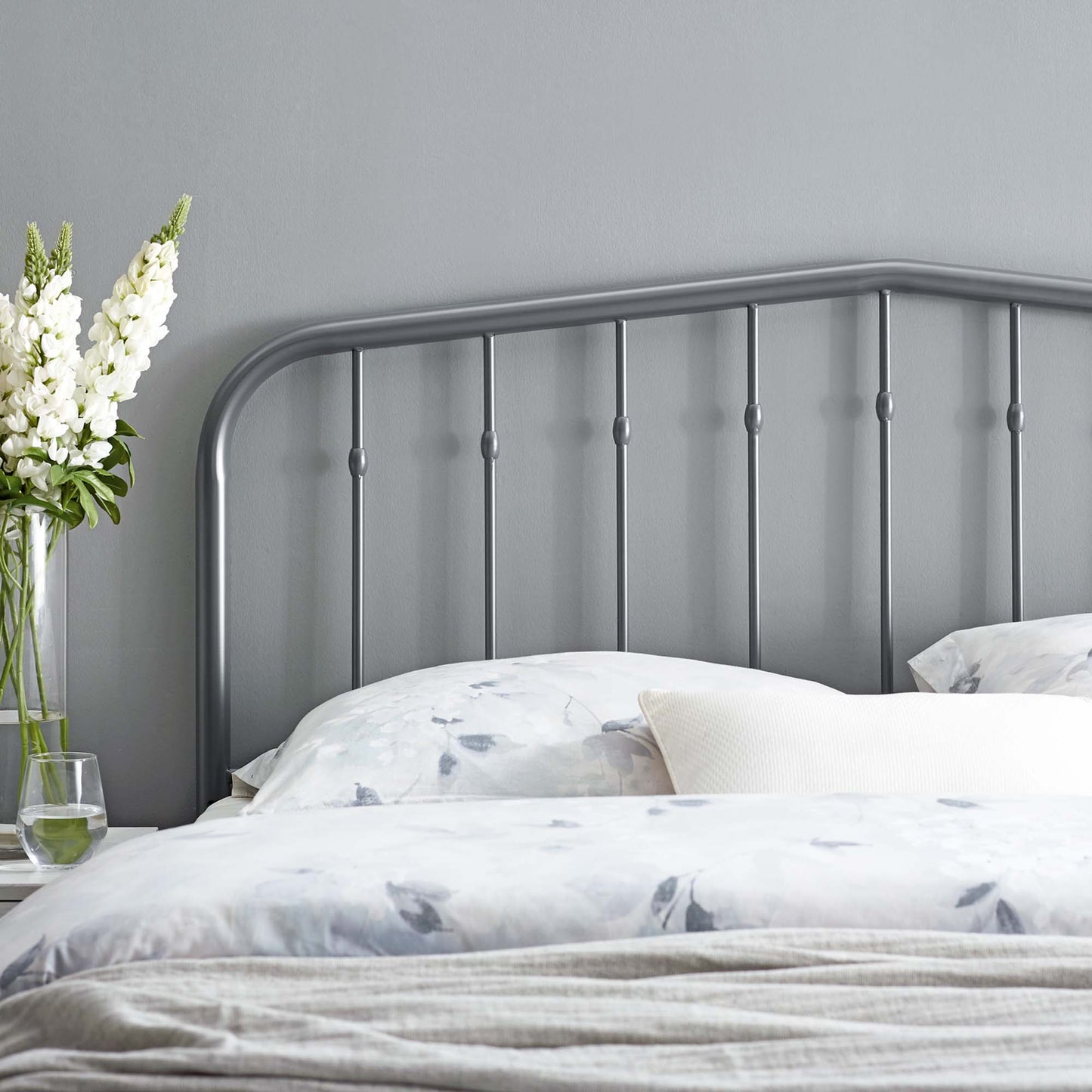 Grey metal headboard with a curved top rail and vertical spindles, placed on light bedding beside a vase of white flowers.