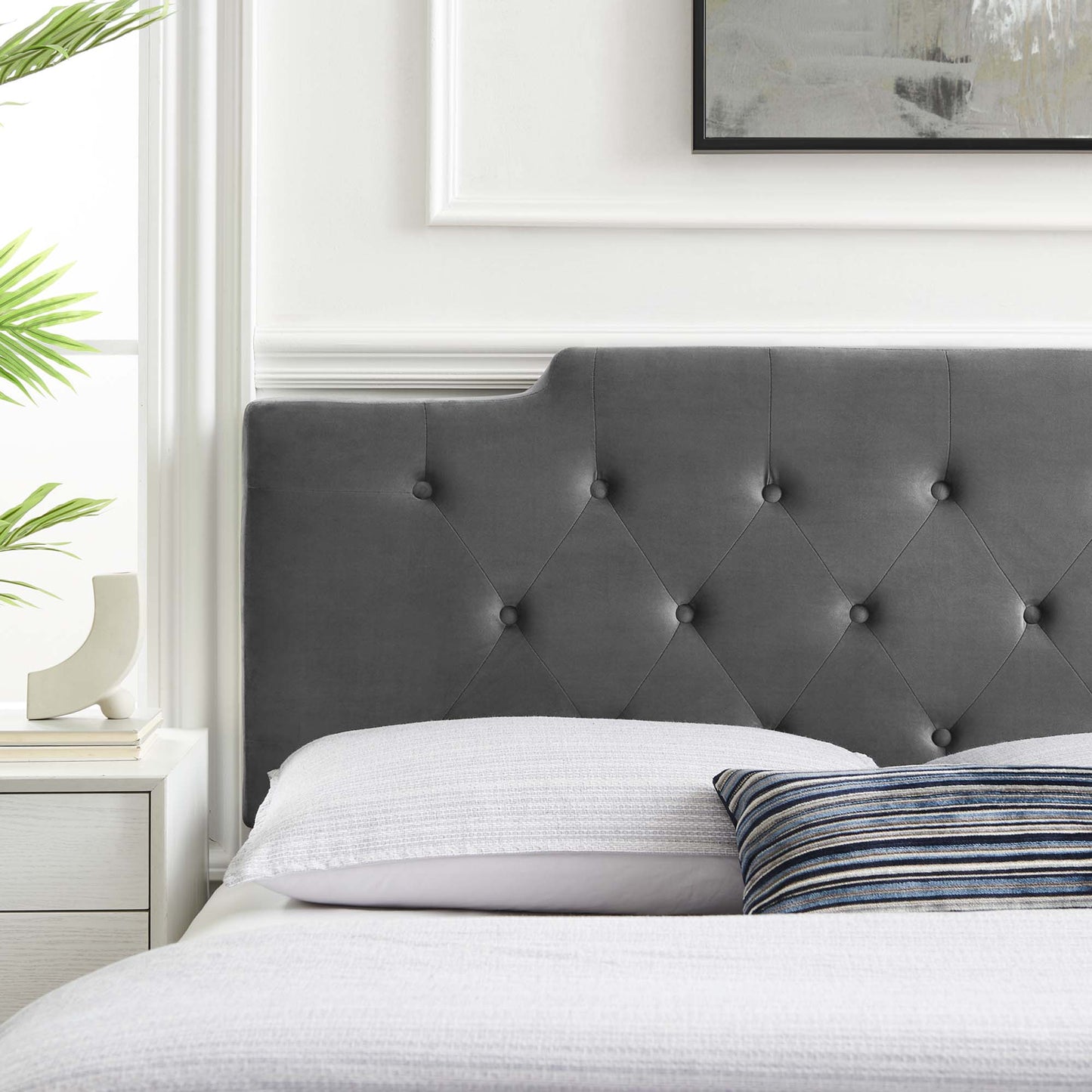 Gray tufted headboard with button tufting in a white-walled bedroom; white bedding & a striped pillow.