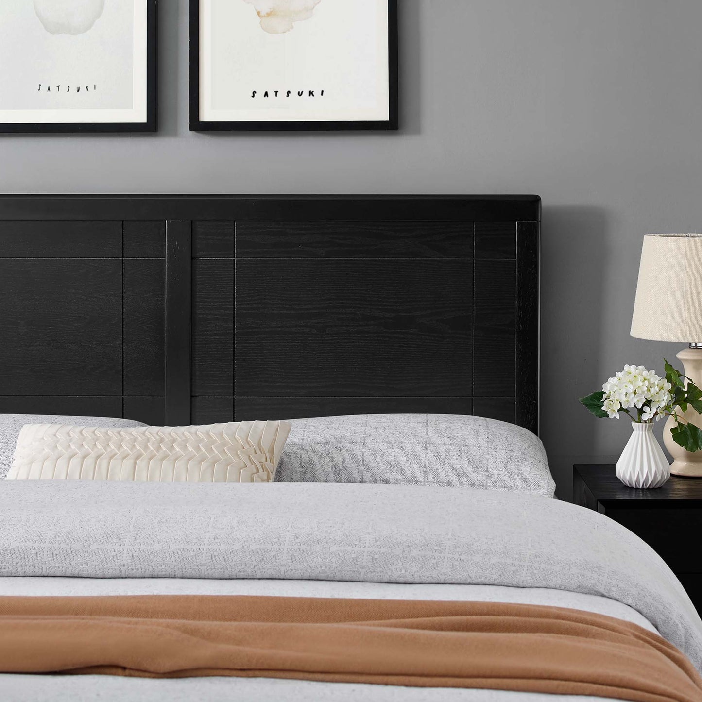 Dark wood headboard, light gray bedding, rust throw, cream pleated pillow; lamp, white flowers, framed abstract prints above.