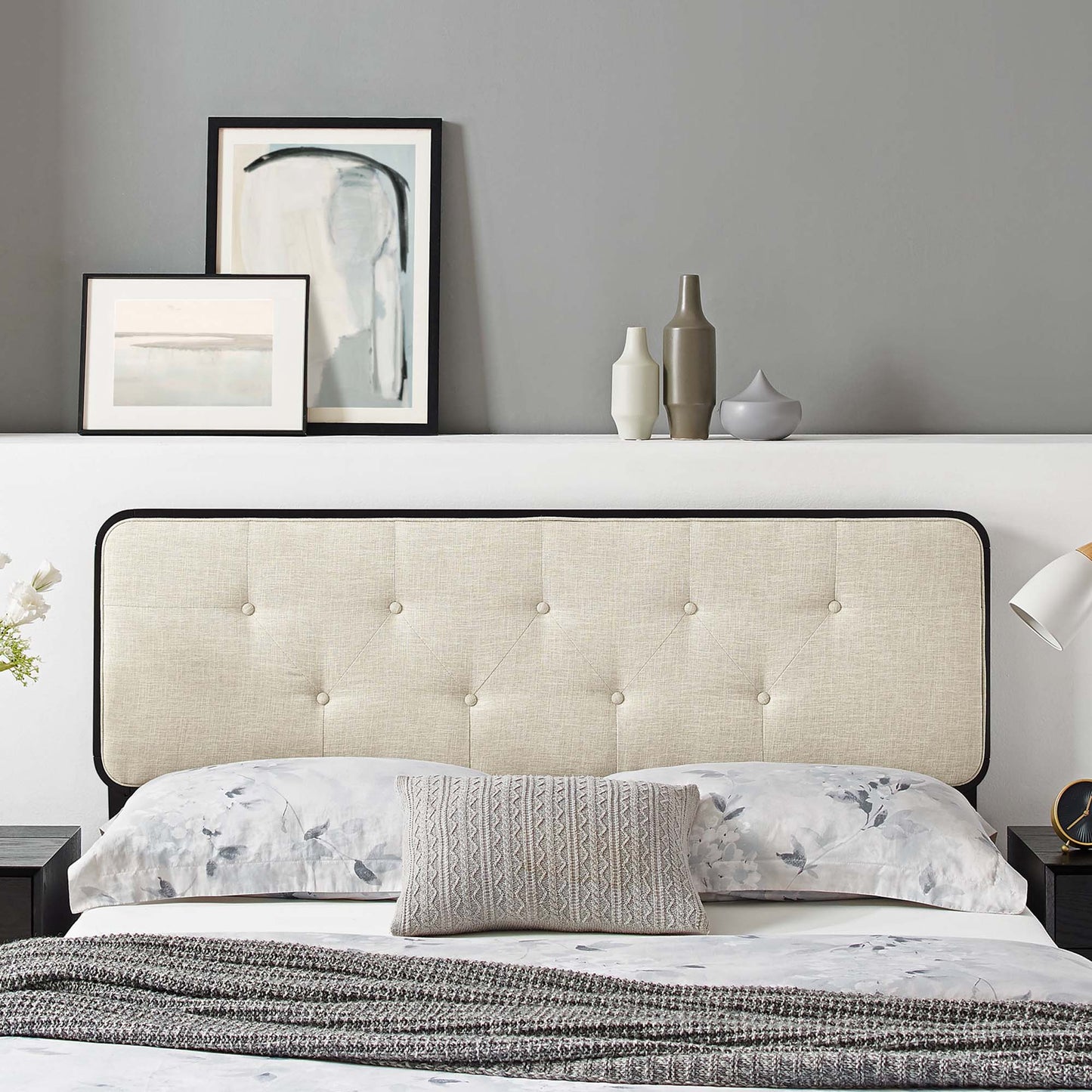 Brown tufted headboard with black trim above a bed with floral bedding, gray knit throw, abstract art and vases on shelf.