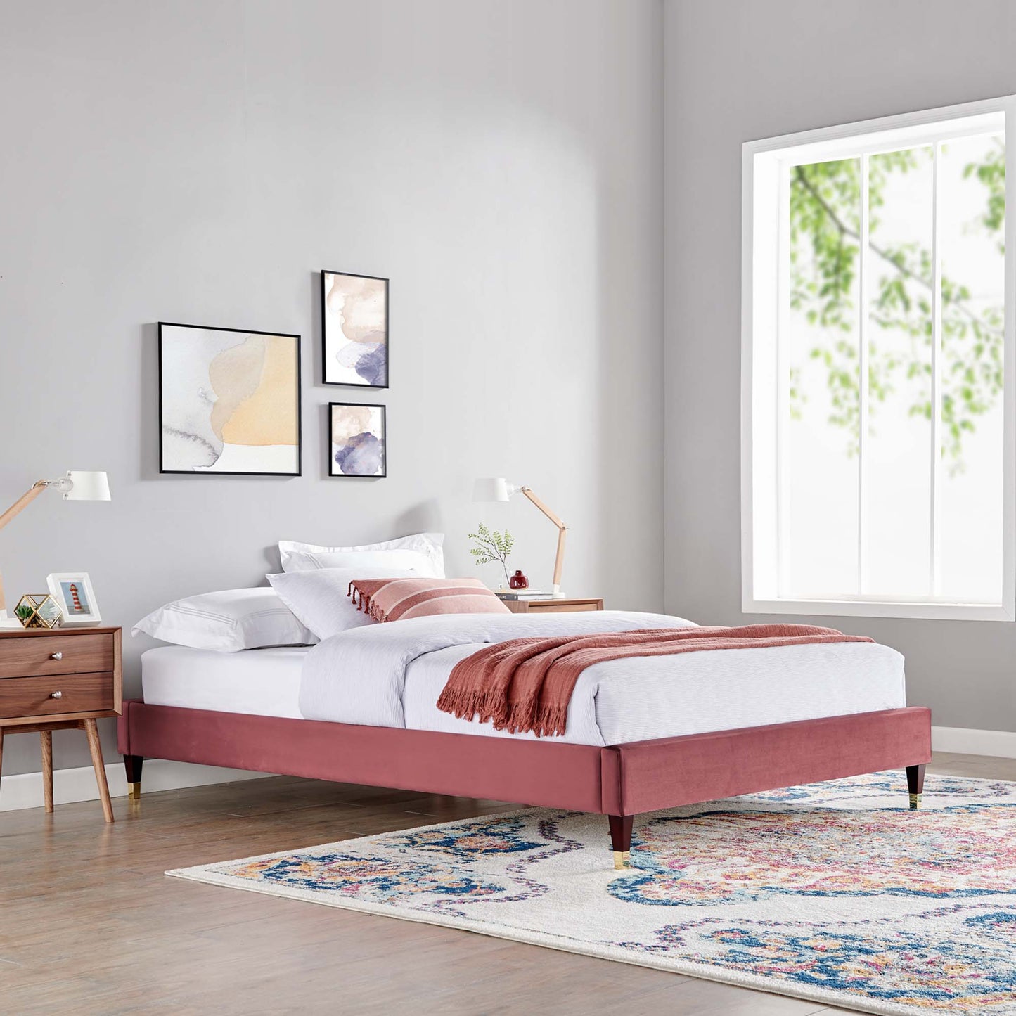 Dusty-rose upholstered platform bed with tapered legs, white linens, pink throw, in a gray modern bedroom with a colorful rug.