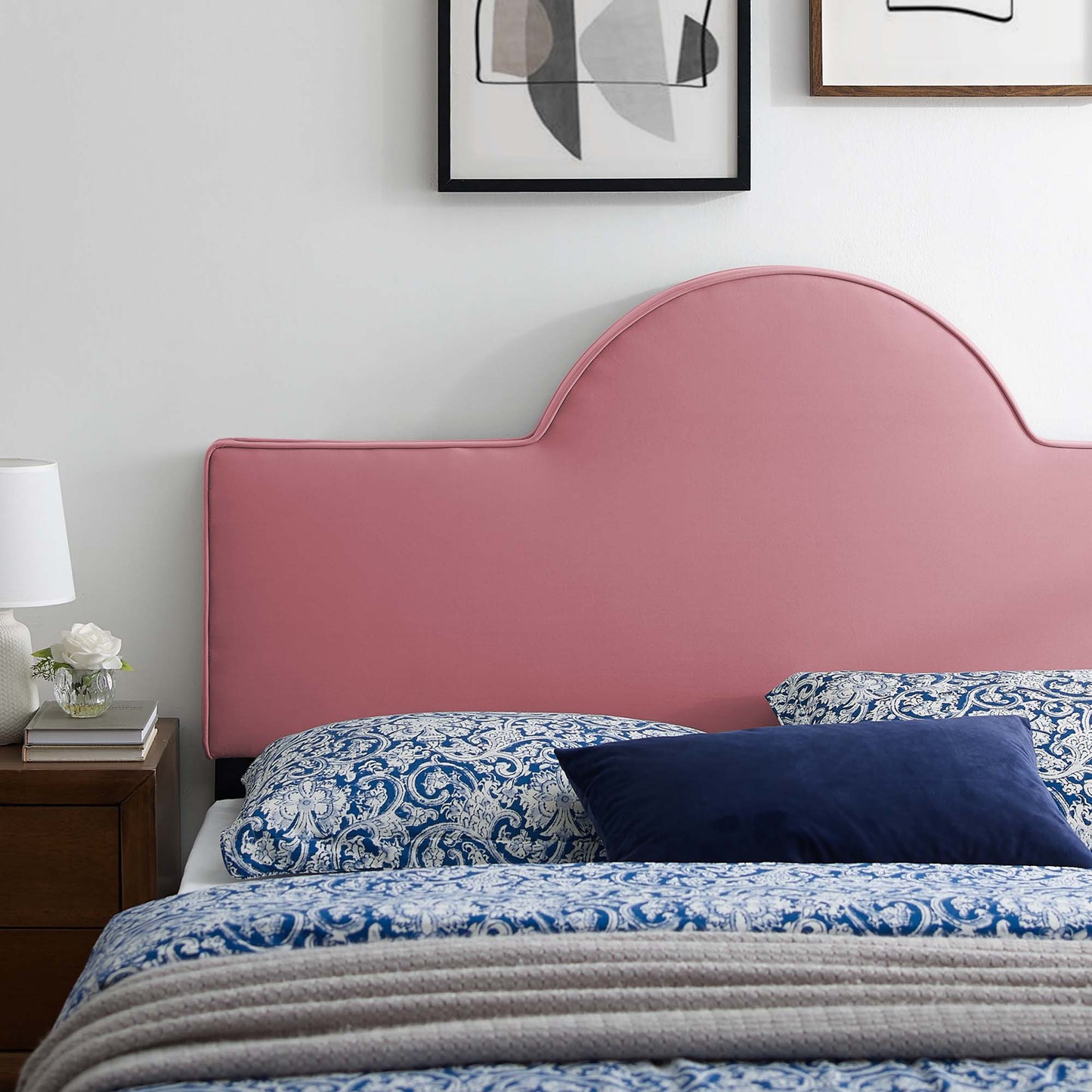 Dusty rose curved upholstered twin headboard with blue paisley bedding and navy throw pillow; wooden nightstand, lamp, framed art.