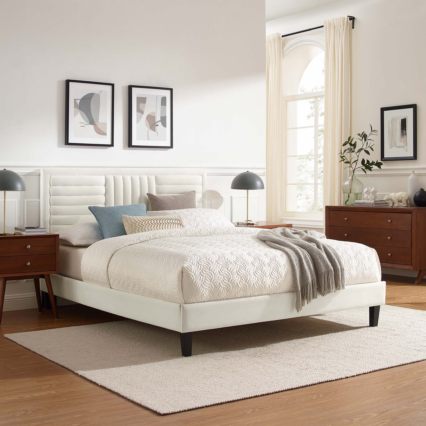 White upholstered platform bed with tufted headboard, cream bedding, gray throw, wood dressers, black lamps, beige rug