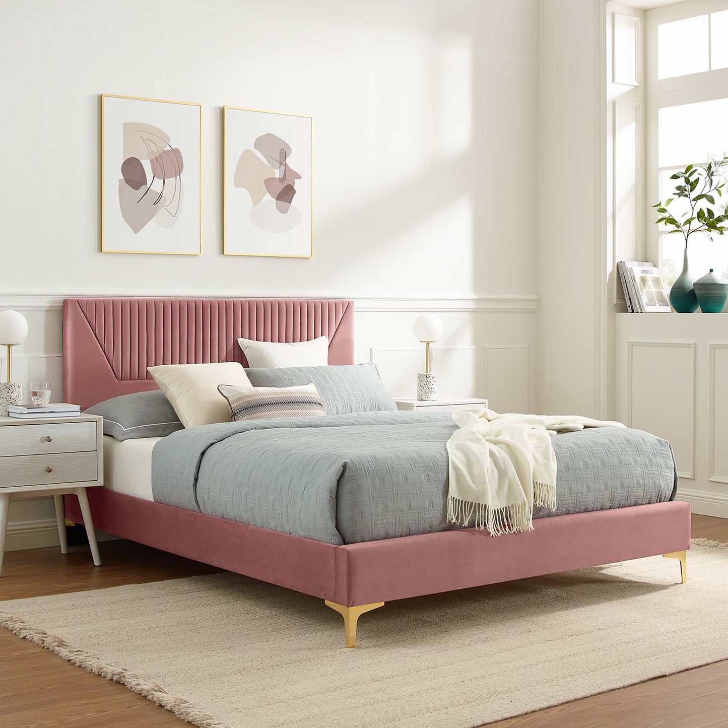 Pink upholstered bed with tufted headboard and gold legs, gray bedding, cream throw, white nightstands, framed abstract art.