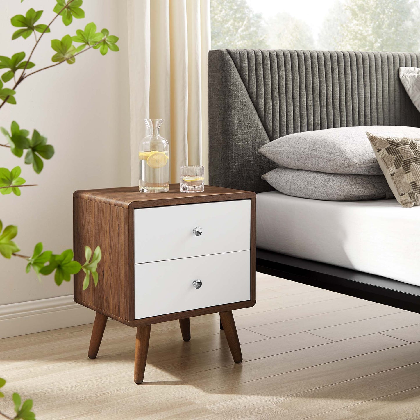 White 2-drawer nightstand with tapered legs beside a gray upholstered bed; glass pitcher on top.