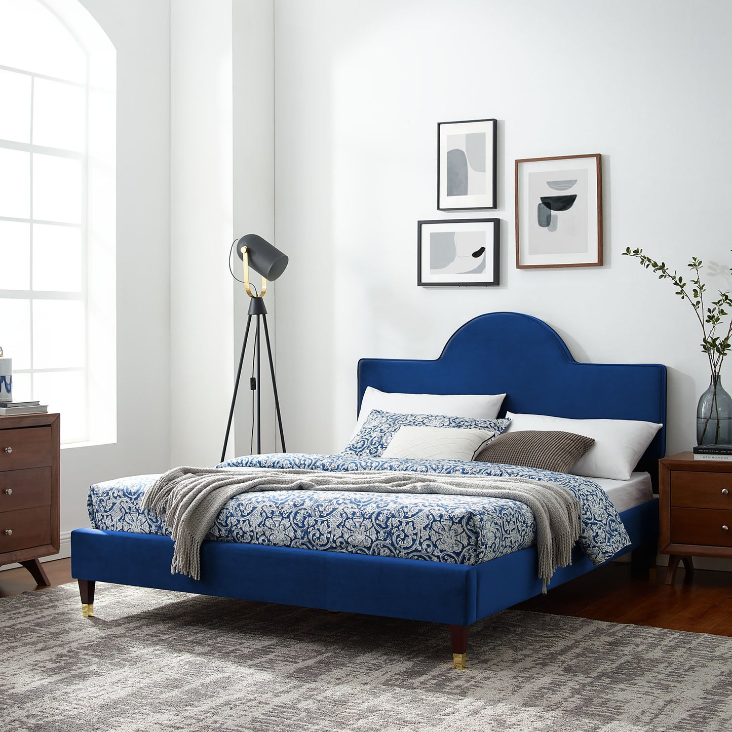 Navy velvet bed with curved headboard, blue-white damask bedding, gray throw, gold-tipped legs; paired with wooden nightstands.