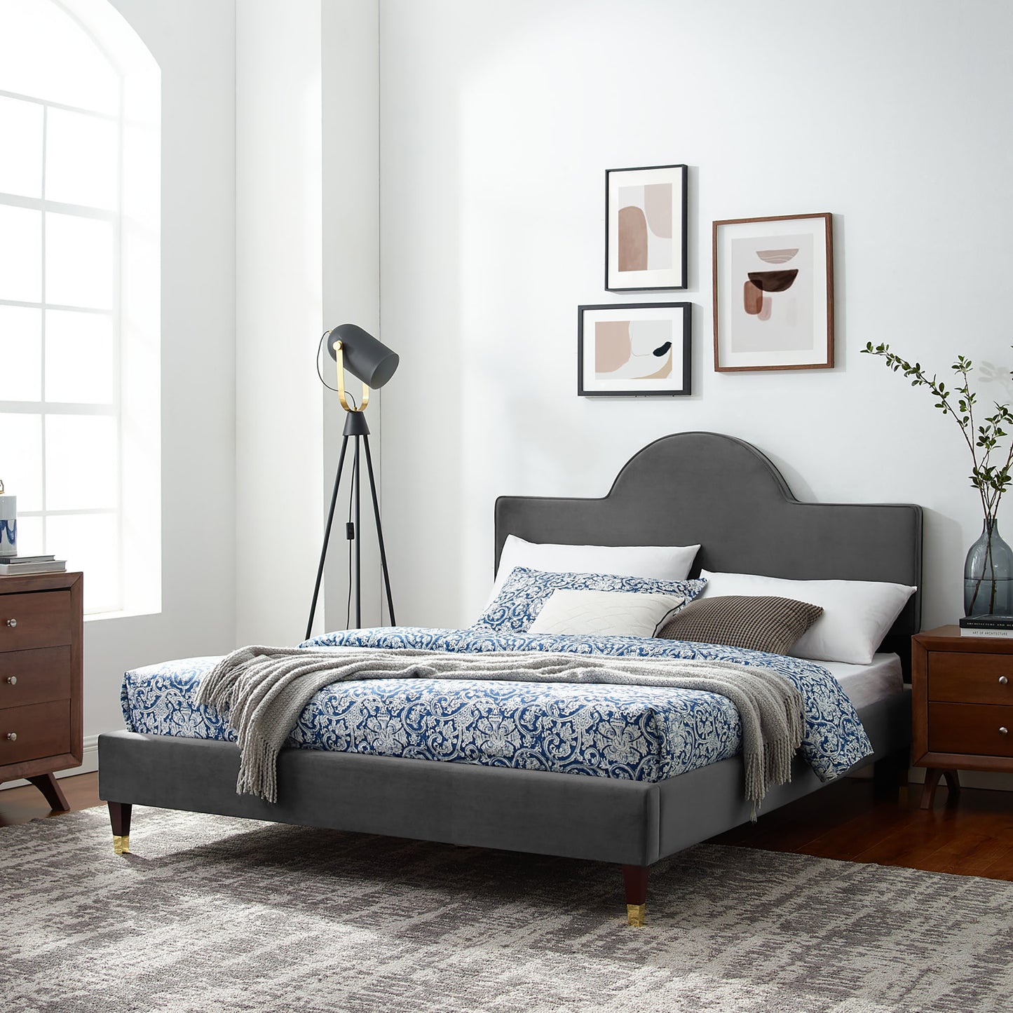 Charcoal upholstered bed with arched headboard, blue damask bedding, knit throw; wood nightstands, tripod lamp, abstract wall art.