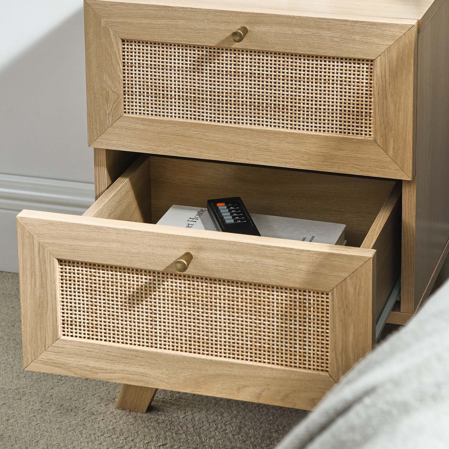 Brown oak wood two-drawer nightstand with woven cane fronts and brass knobs; bottom drawer open with remote inside.