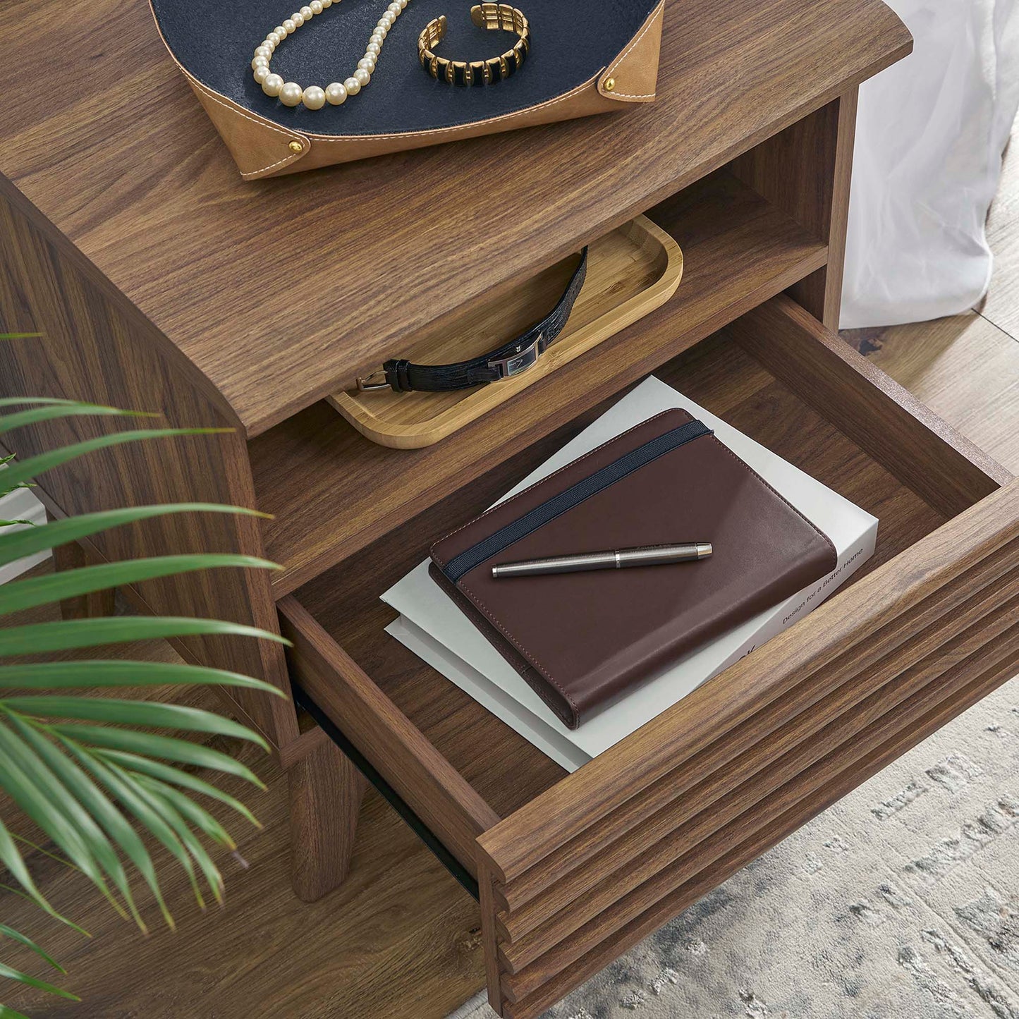 Walnut wood nightstand with drawer & top tray; brown leather wallet & pen in the lower drawer, pearl jewelry on the top tray, plant nearby.