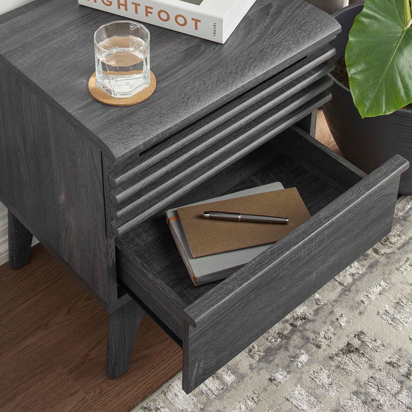Dark gray wood nightstand with grooved sides; open drawer with notebook and pen; glass of water on a cork coaster.