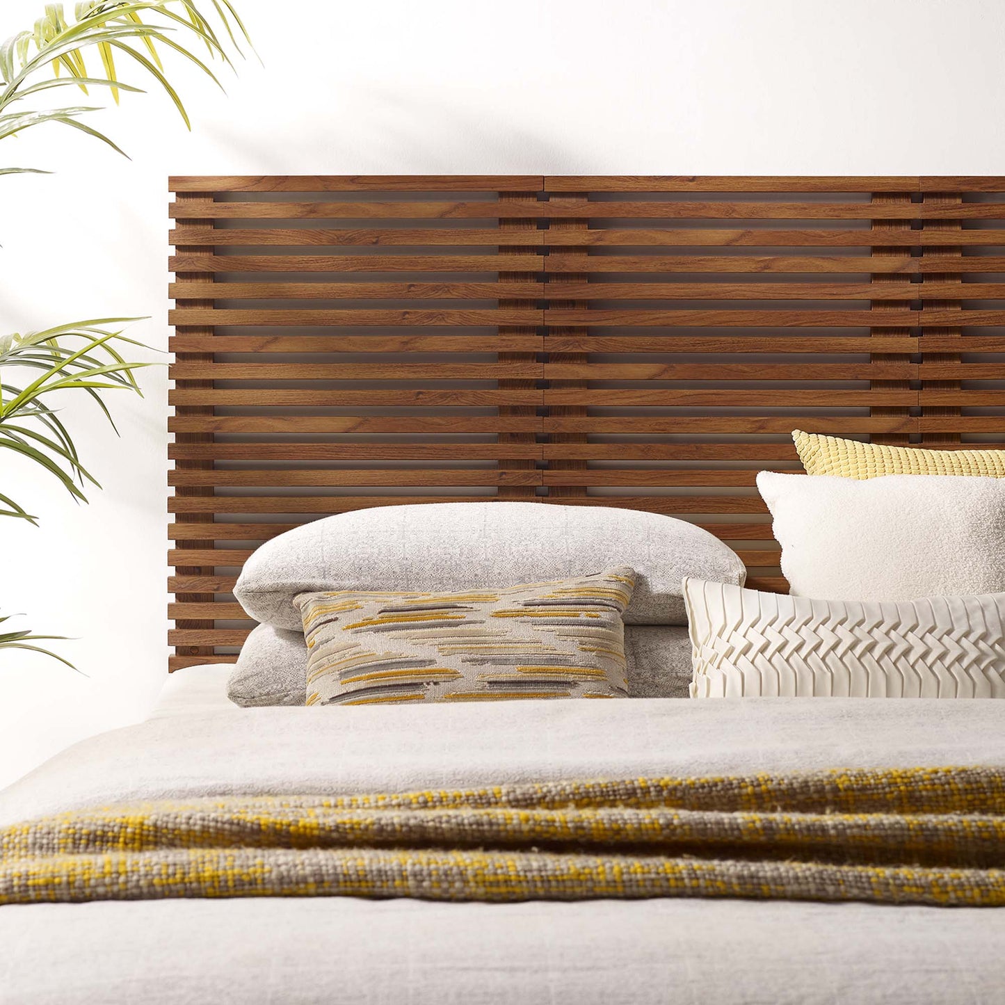 Brown, wall-mounted queen headboard with a wooden slat design; neutral gray linens, striped and chevron pillows, mustard accents; plant nearby.